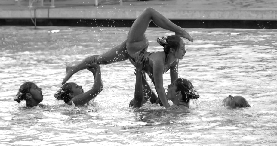 A group of children is practicing synchronized swimming in a pool, with one girl performing a handstand while others support her.