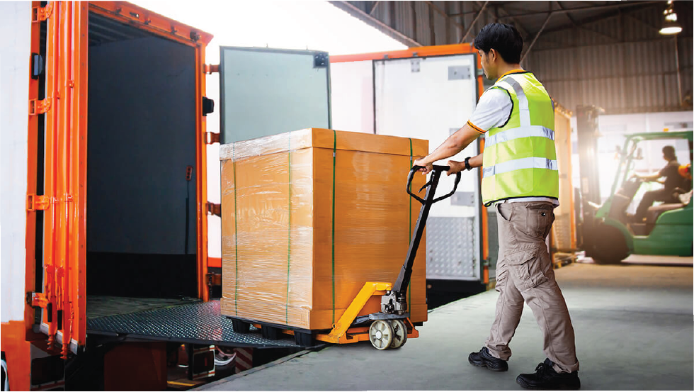 A worker in a safety vest pushes a pallet of boxes on a hand truck into a truck in a warehouse, with a forklift in the background.