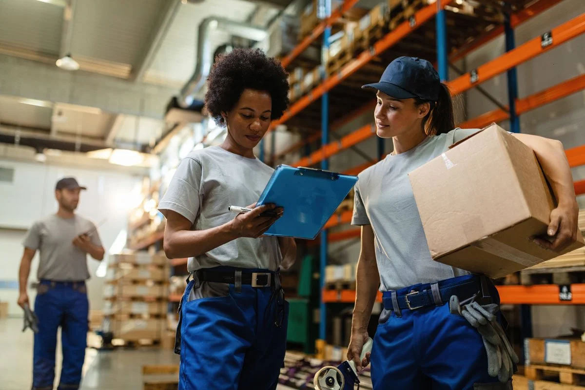 Three warehouse workers in a storage facility, two women in the foreground reviewing a clipboard and holding a box, and a man in the background using a mobile device.