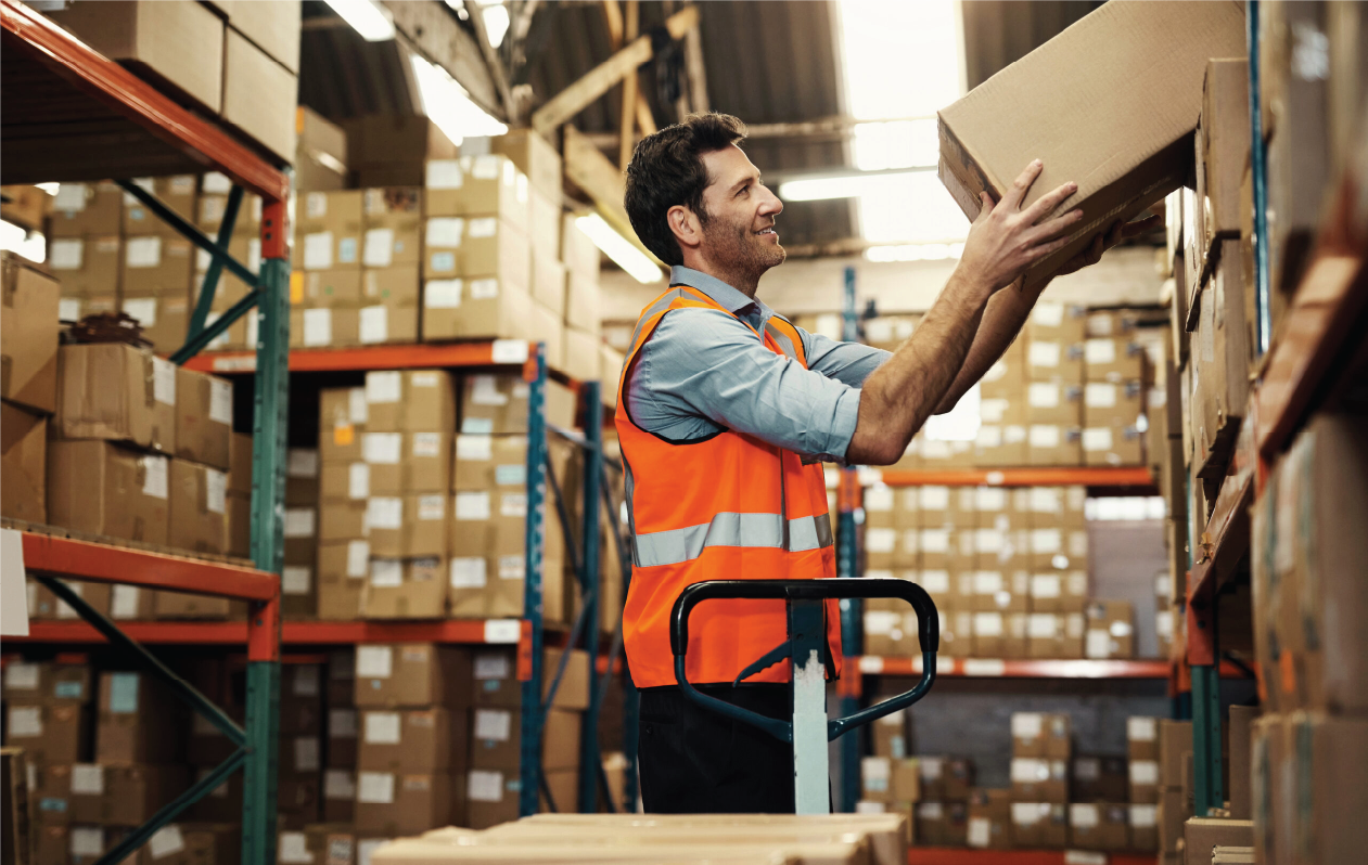 A man in a warehouse wearing an orange safety vest, lifting a cardboard box while standing next to a cart, surrounded by shelves stocked with numerous boxes.