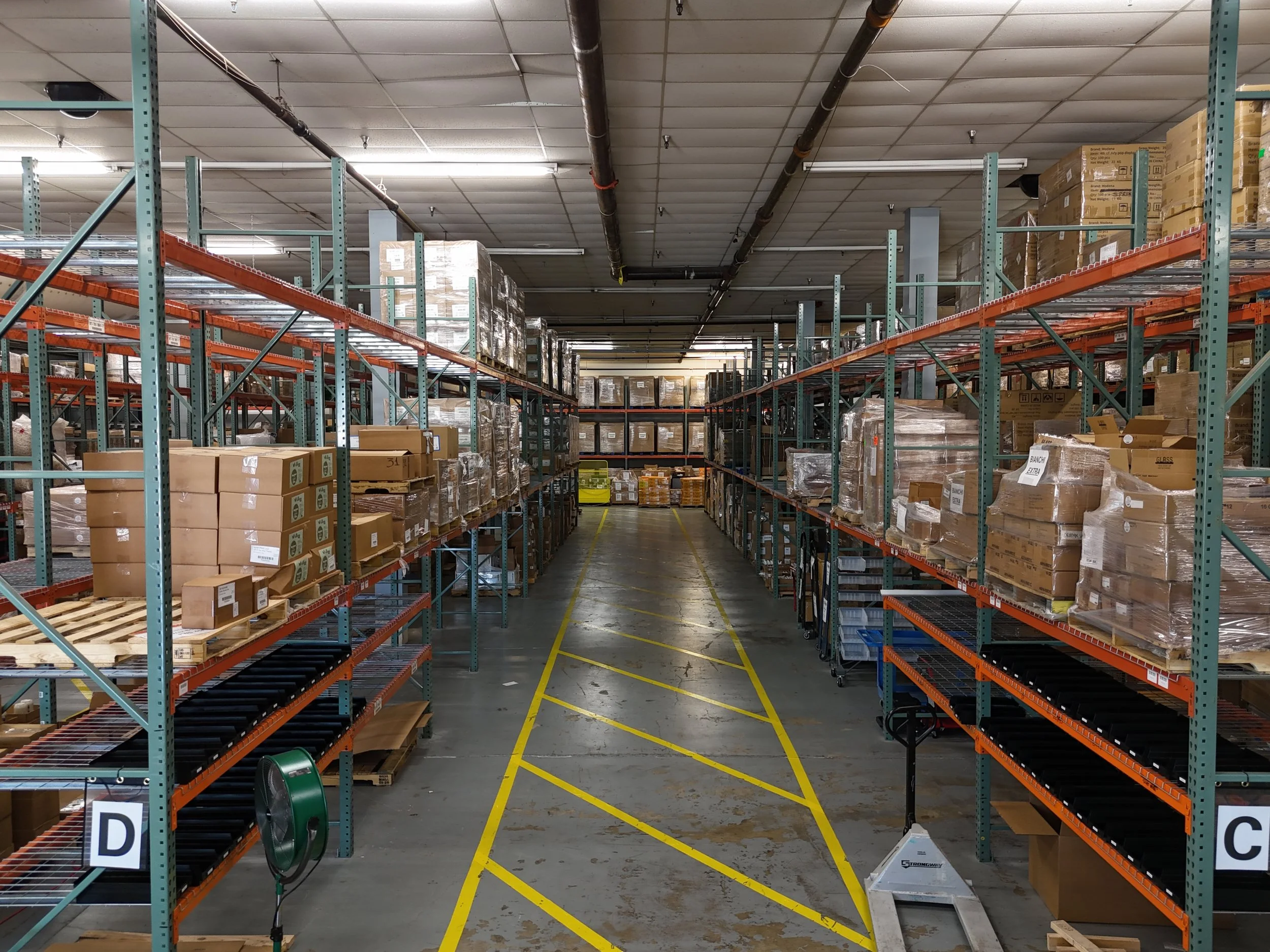 Warehouse aisle with metal shelving filled with cardboard boxes, some wrapped in plastic, stacked on both sides, yellow safety lines and labels on the floor, fluorescent lighting, ceiling pipes.