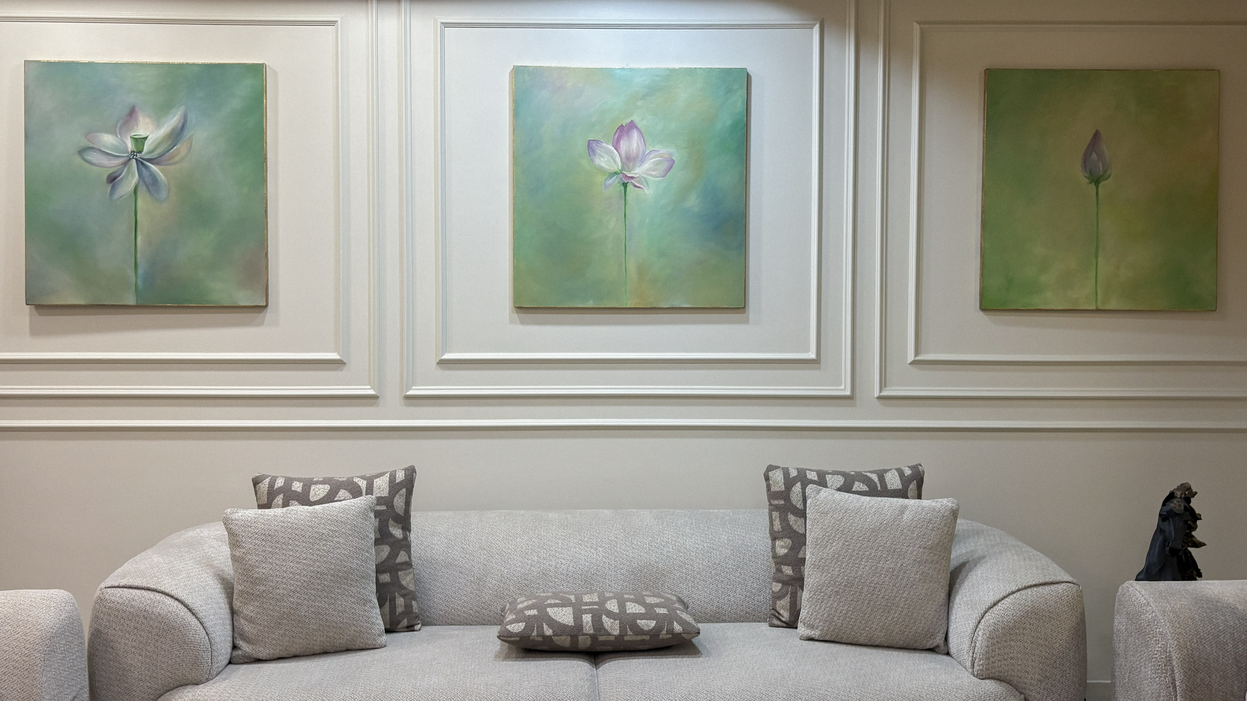 Living room with beige sofa, patterned throw pillows, and a framed art arrangement of three vertical floral paintings with pastel tones on a white wall.