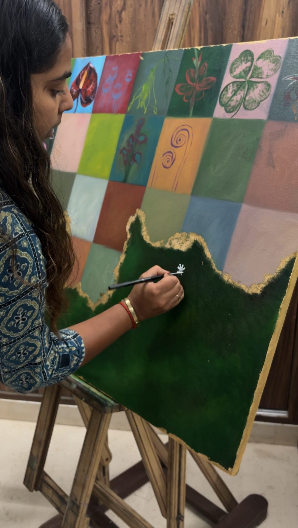A woman painting a landscape on a large canvas that is propped on a wooden easel, featuring a green mountain and a small white flower.