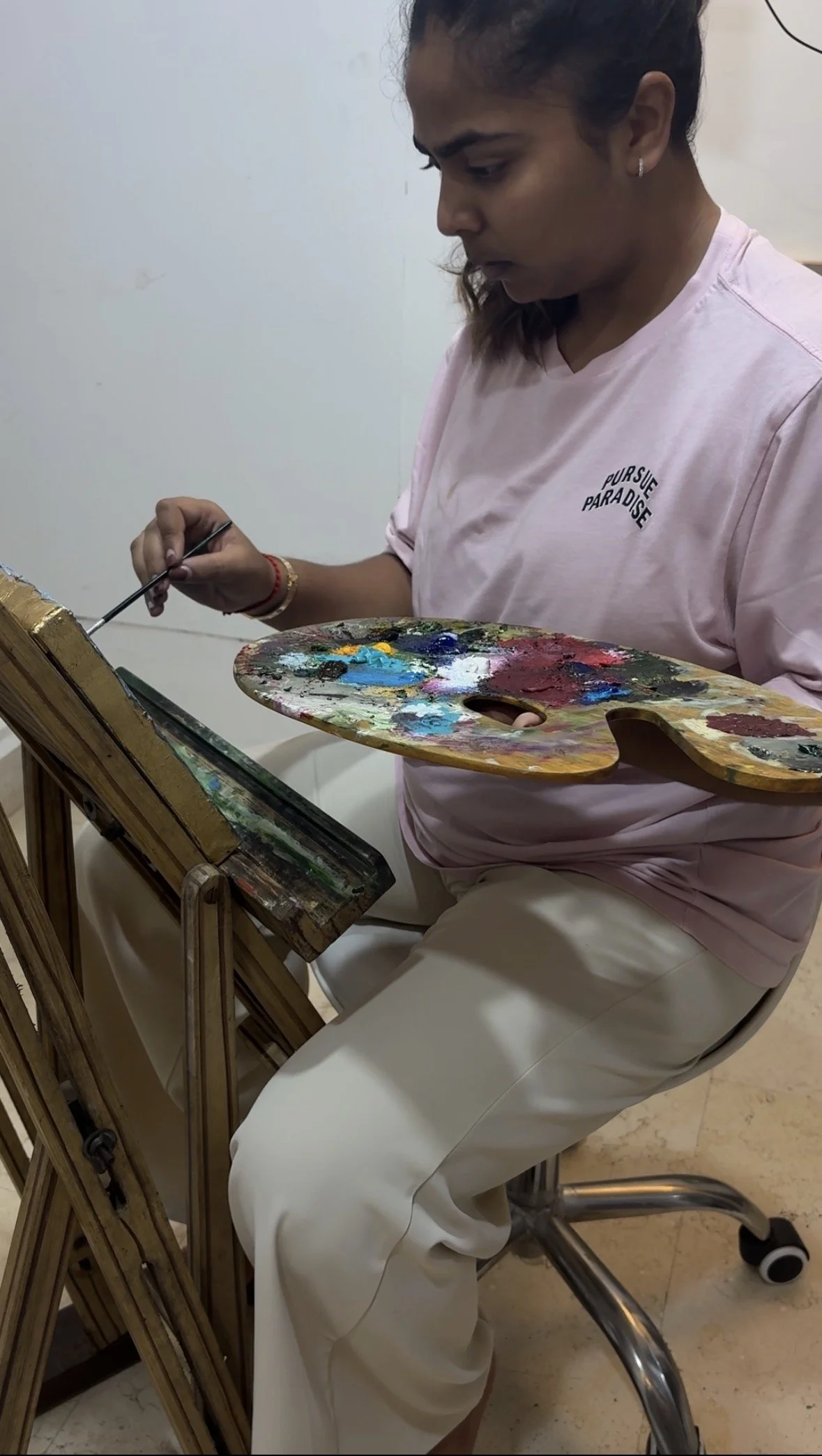 A woman is sitting on a rolling chair, painting on a canvas, holding a palette with various colors in her left hand and a brush in her right hand. She is focused on her artwork, wearing a pink T-shirt with the text 'Pursue Paradise' and white pants.
