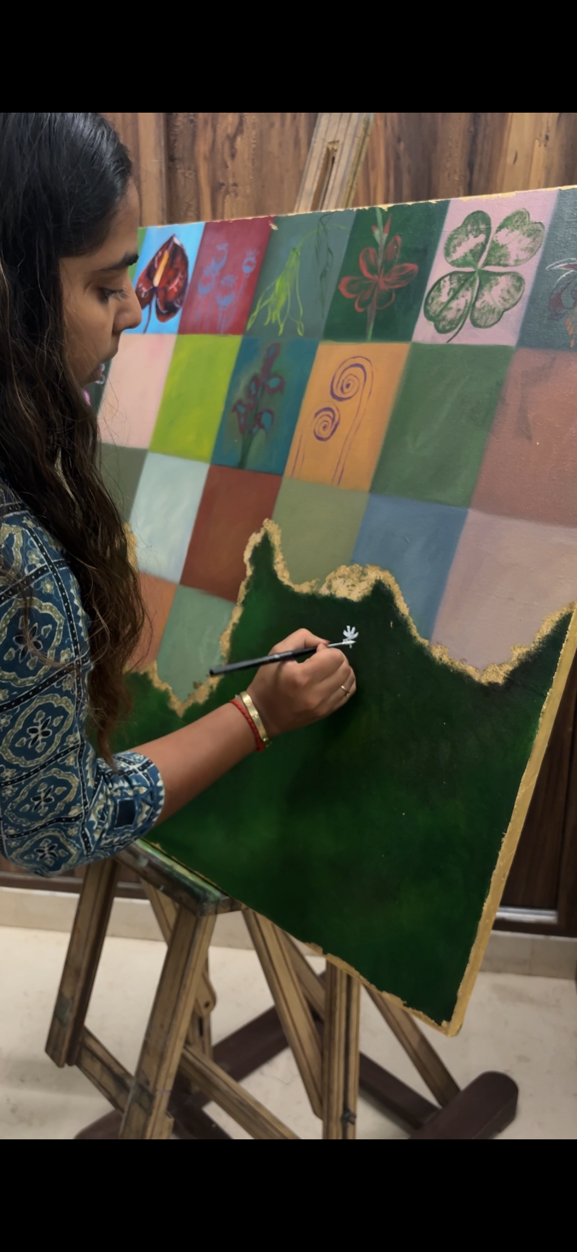 Woman painting on a colorful canvas featuring flower designs and sections of solid colors. She is using a small brush to add details to a green section of the painting, standing in front of a wooden easel.