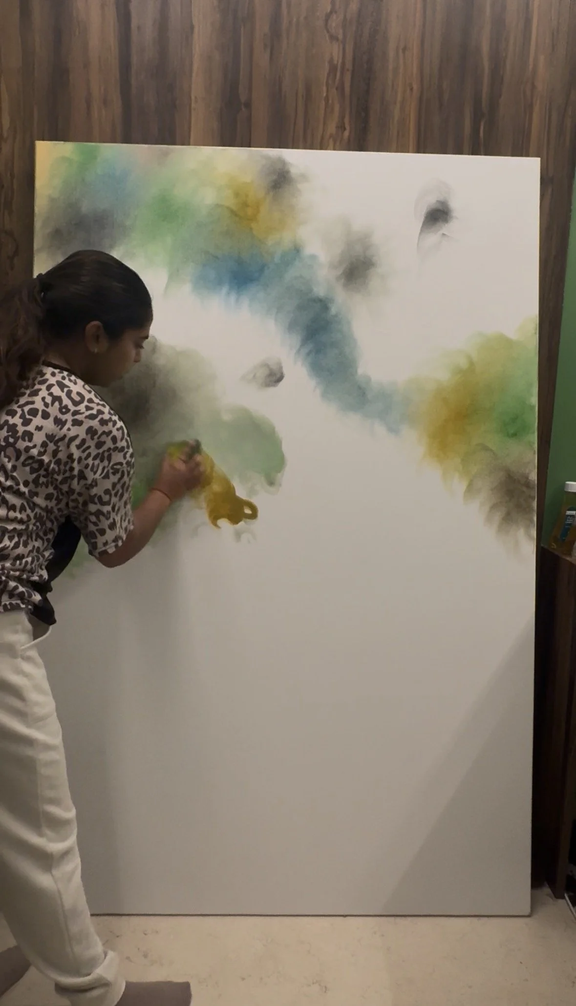 A young girl with a ponytail and leopard print shirt is painting with yellow, green, blue, gray, and black watercolor on a large white canvas in front of a wooden wall.