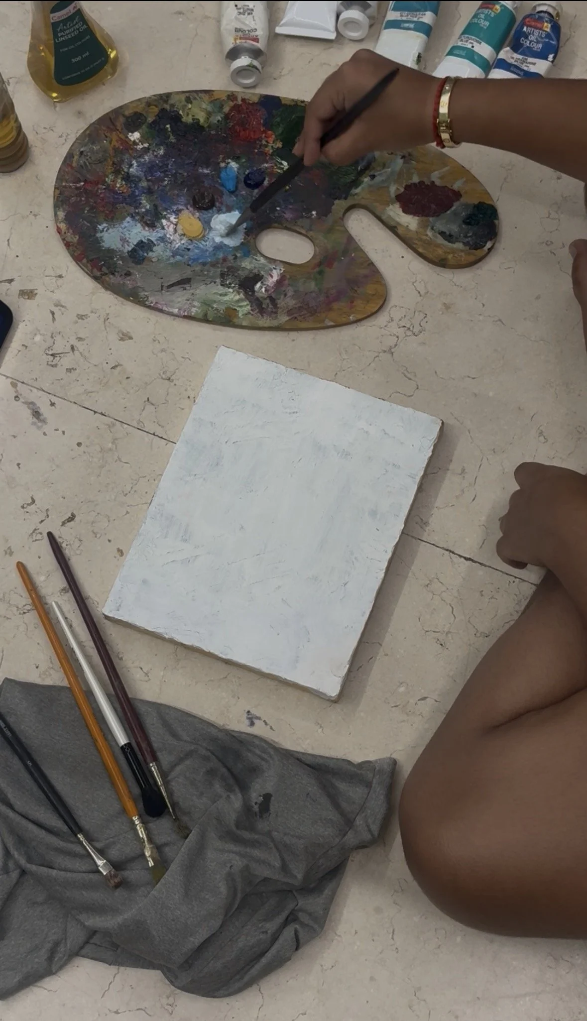 Person painting a white canvas with a paintbrush, surrounded by paint tubes, a palette with mixed colors, and pencils on a marble surface.
