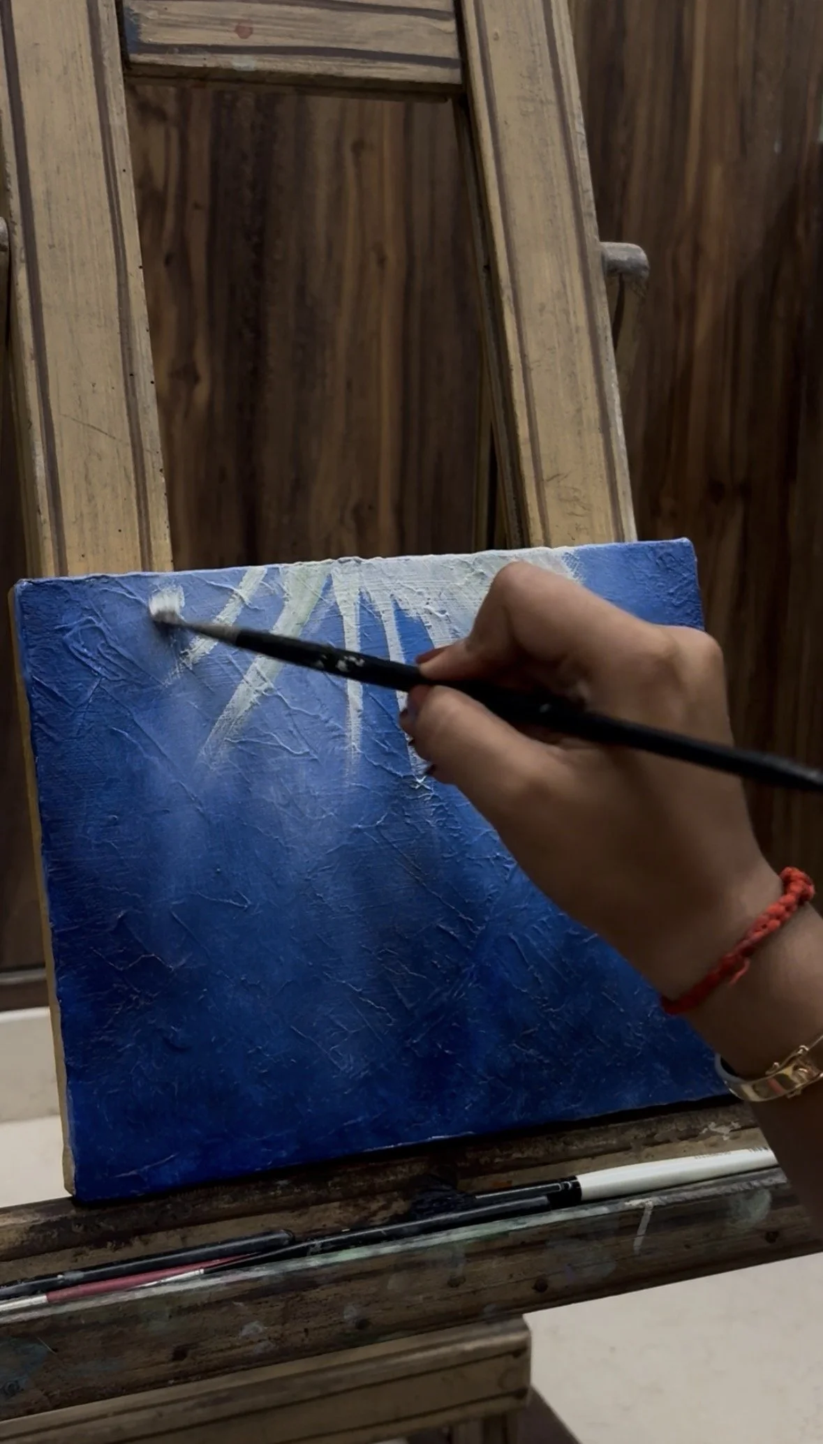A person is painting a small canvas with blue and white abstract strokes using a fine brush, on a wooden table with various brushes and tools around.
