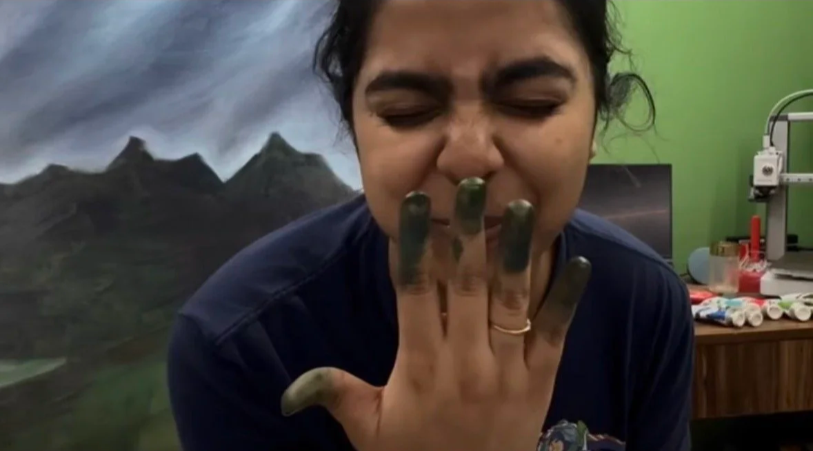 Person with dark hair and a ring on finger, covering nose with dirty hand, in a room with green wall and mountains painting in the background.