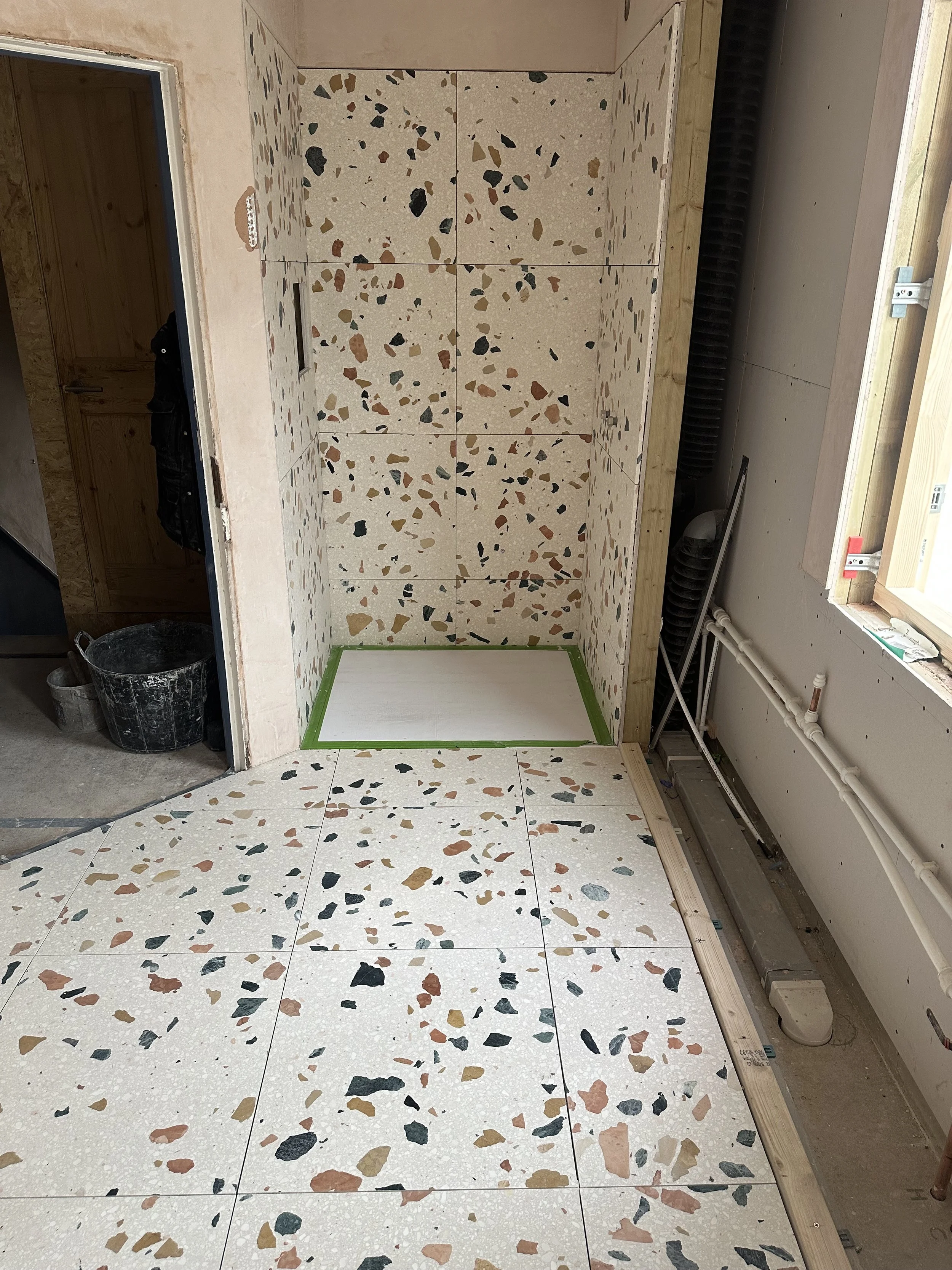 Interior of a room under construction or renovation with terrazzo flooring and walls, exposed pipes, and a window with natural light.