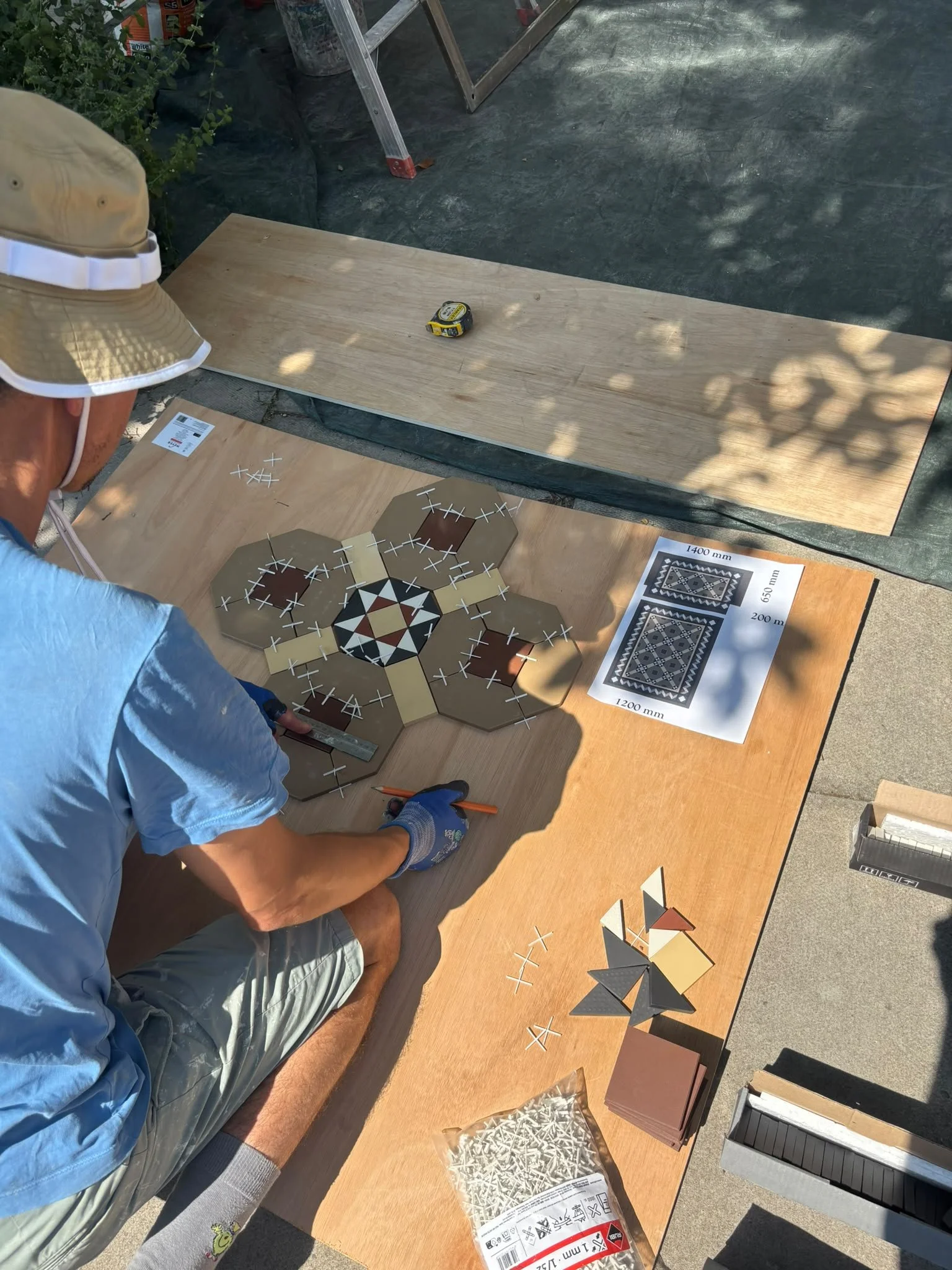 A person working on a woodworking or craft project at a table outdoors. The person is wearing a beige bucket hat, a light blue shirt, and gloves. The table has various tiles, some with hexagonal patterns, as well as a sheet with different tile sizes 