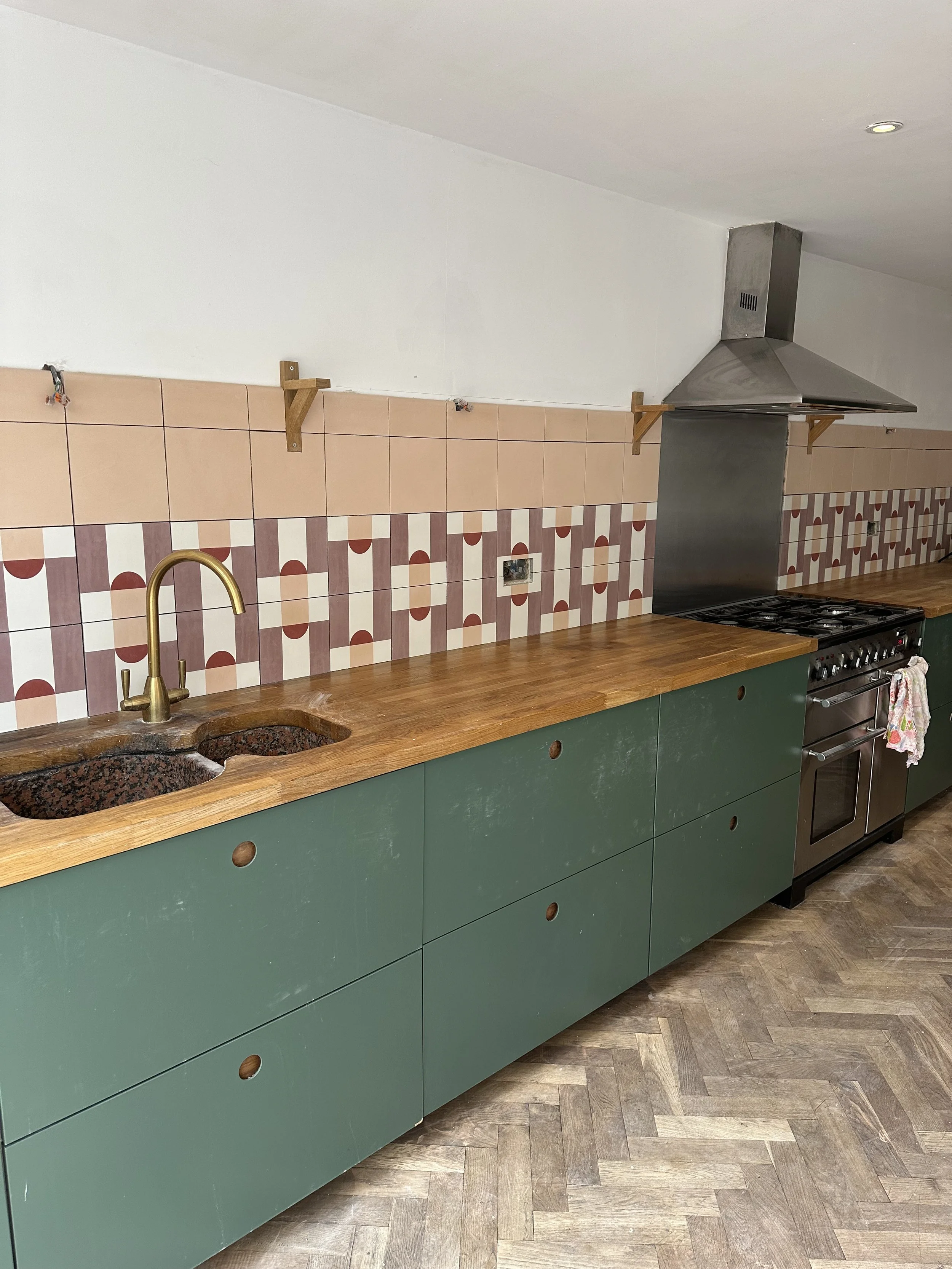 Empty kitchen with green cabinets, wooden countertop, tiled backsplash with geometric pattern, stainless steel stove, and hanging wooden supports.