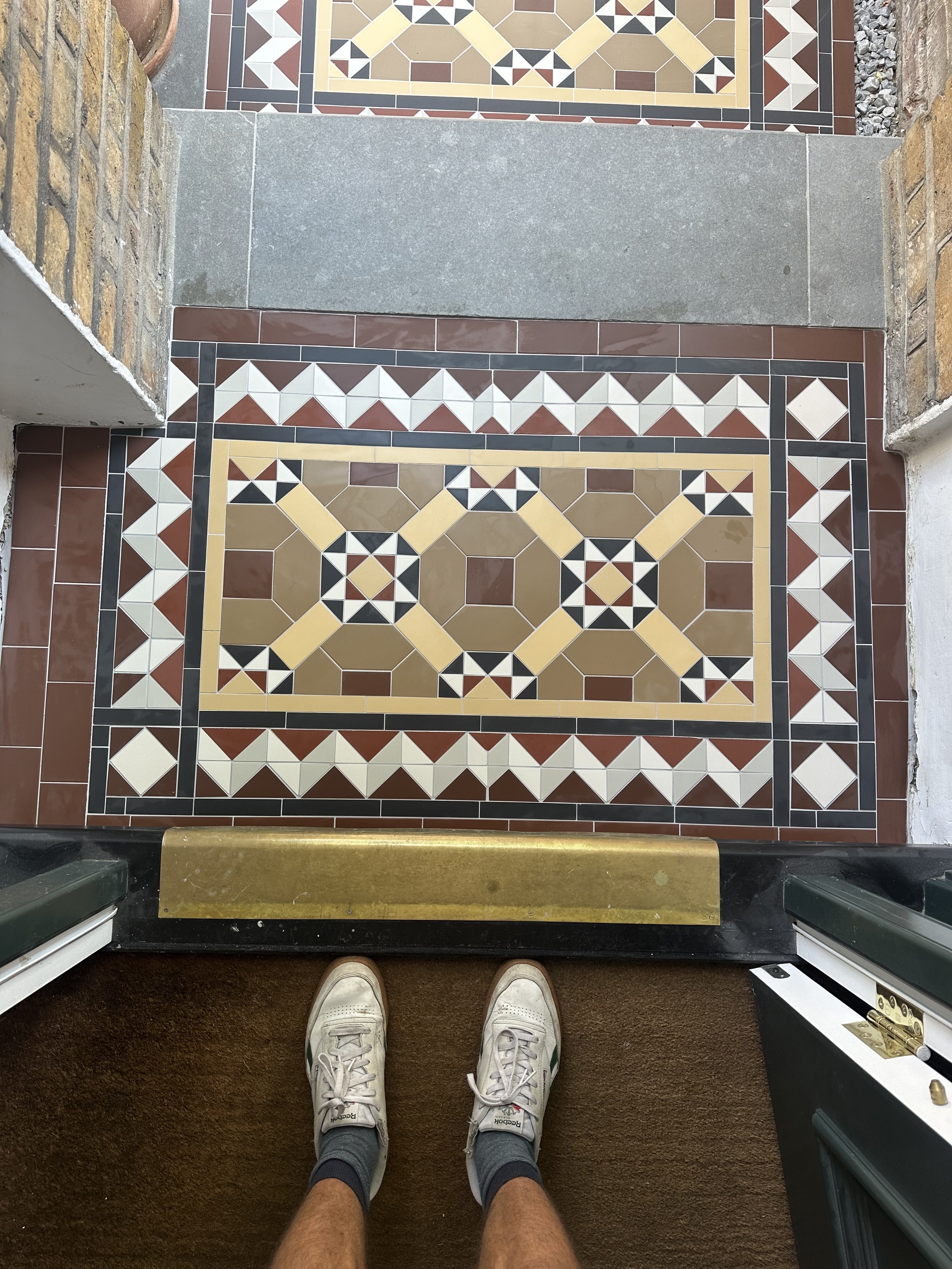 Person standing at the threshold of a tiled floor, wearing white sneakers, with the camera positioned at a downward angle.