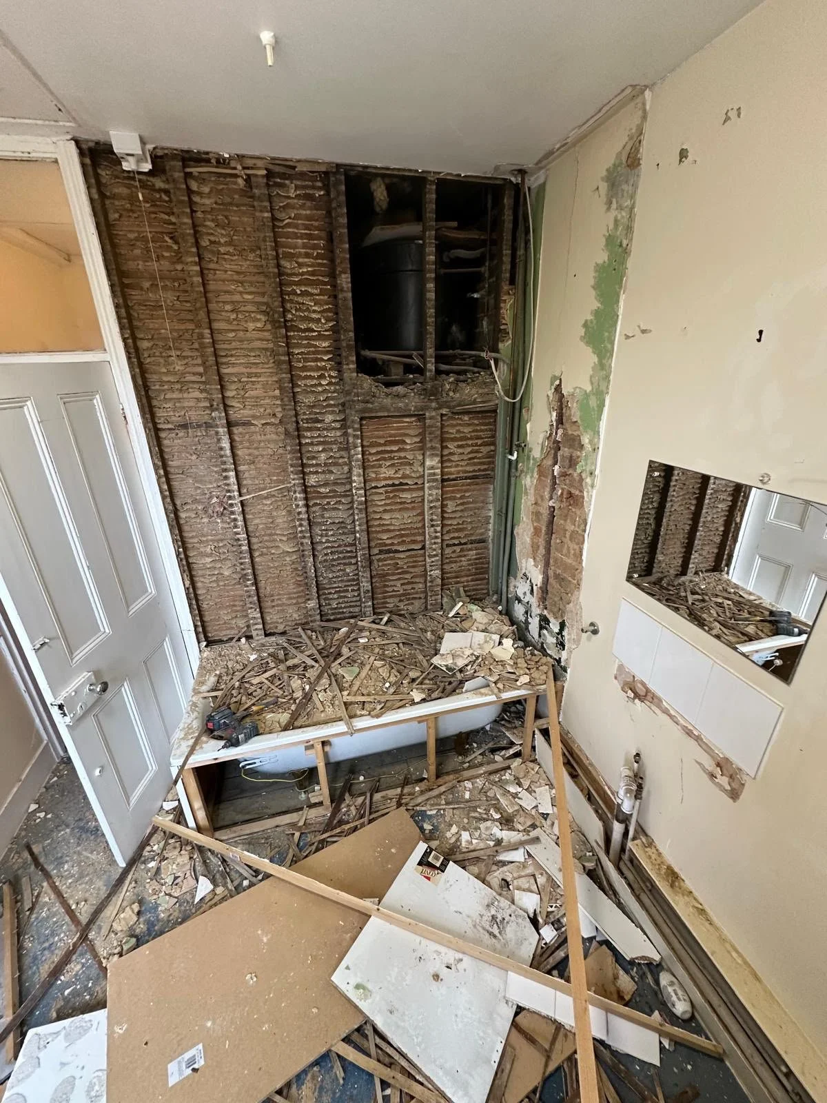 Room under renovation with exposed wall studs, debris on the floor, a partially removed bathtub, a mirror, and an open door leading to another room.