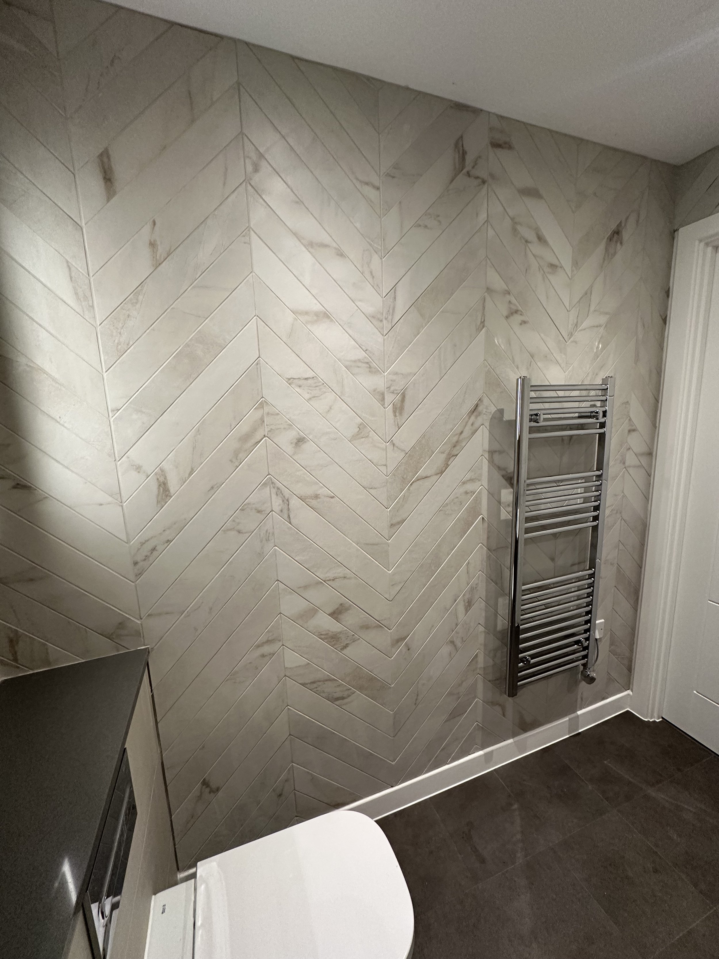 A restroom corner with beige and white marbled tile walls, a stainless steel heated towel rack, a small section of a toilet, a small black countertop, and dark flooring.