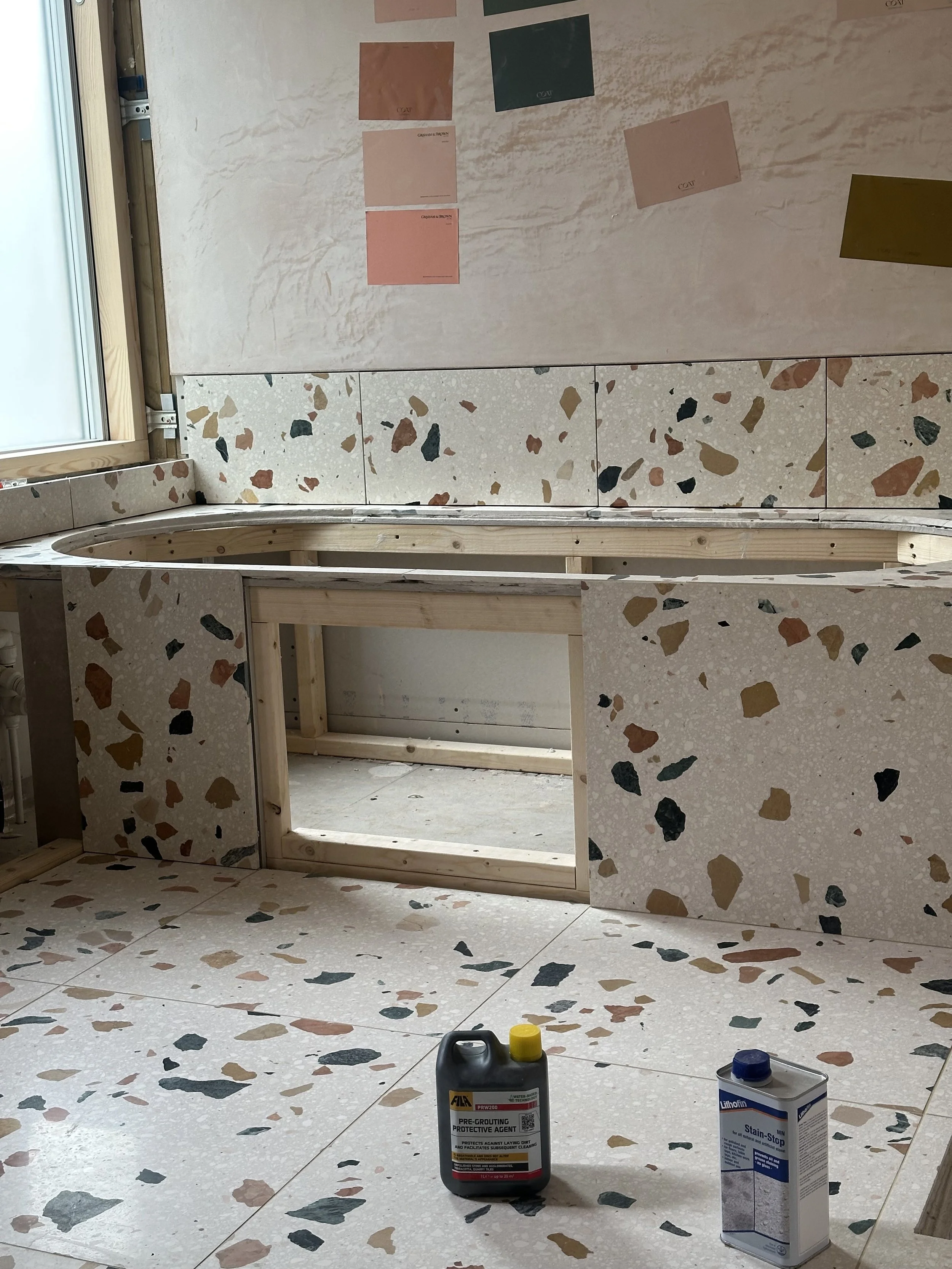 Interior of a building under construction with terrazzo tile flooring and wall panels. There are wooden frames and placement of tiles, with bottles of grout or sealant on the floor, and a window on the left side.
