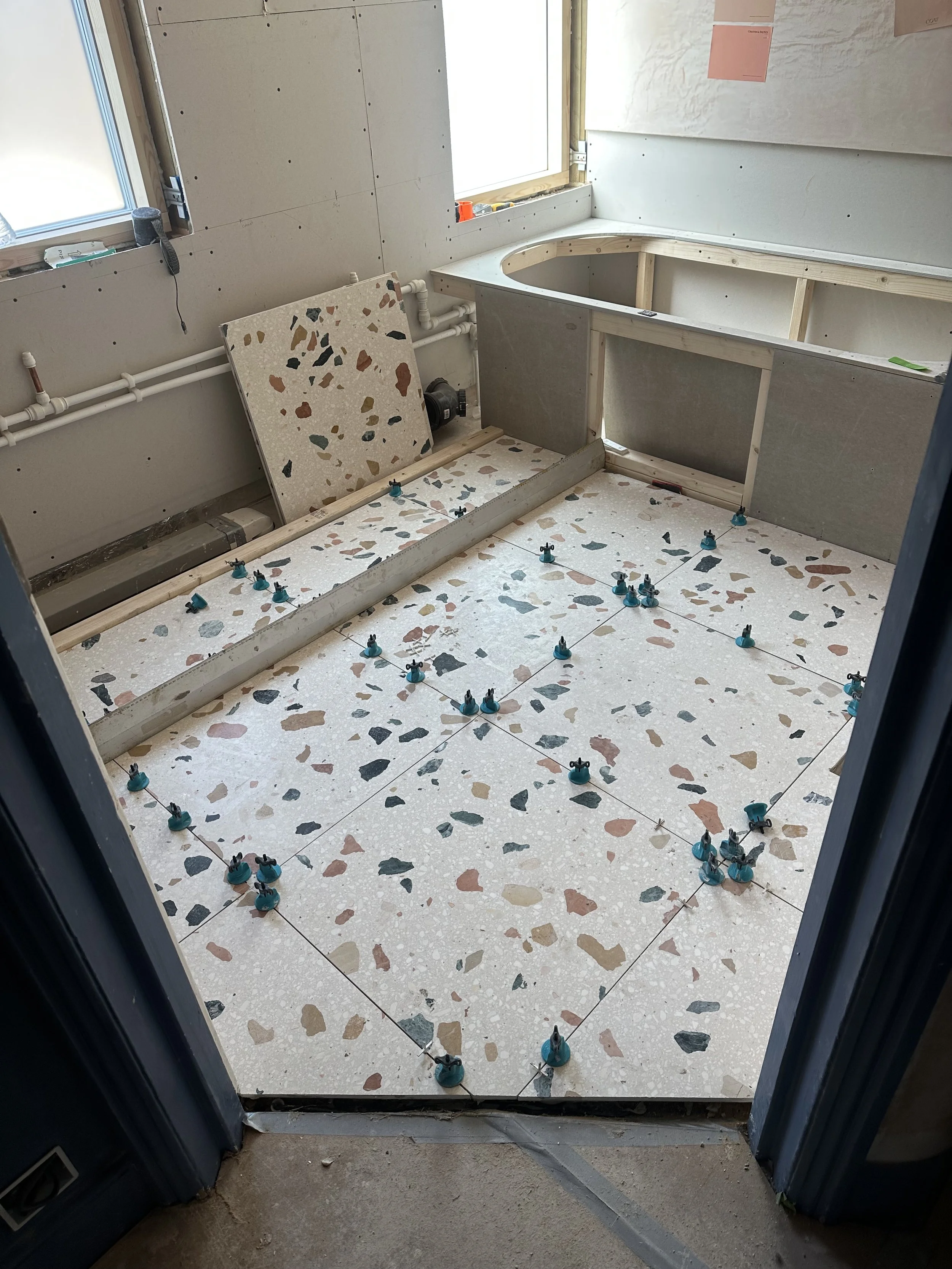 Bathroom under construction with terrazzo tile flooring, a tub installation in progress, and uninstalled cabinetry. Bullet anchors are placed on the floor, and plumbing pipes are visible behind the tub. Two windows are in the background, one with a c