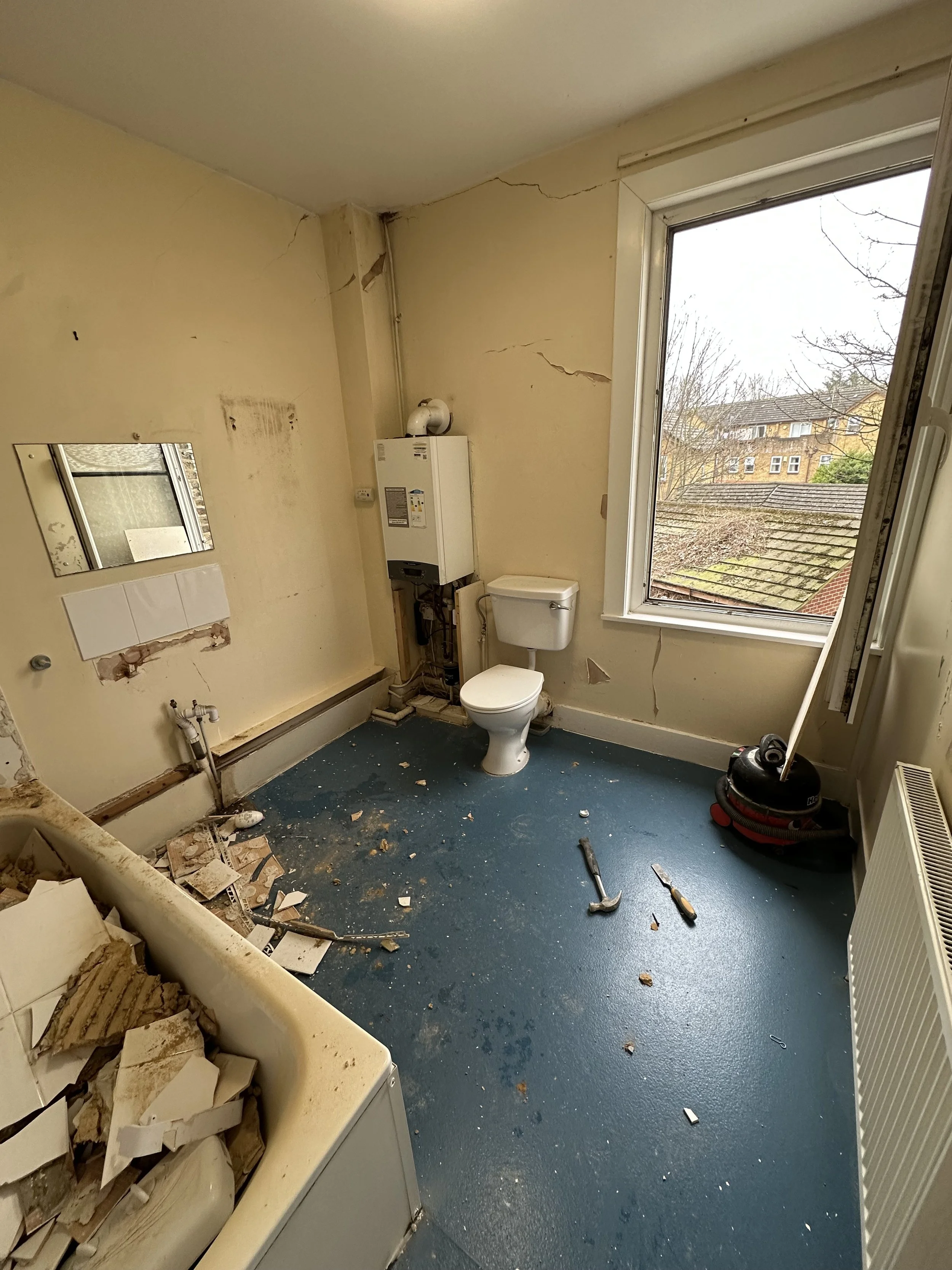A bathroom with damaged walls and ceiling, cracked walls, debris on the floor, an open bathtub filled with debris, a toilet, a small mirror, and tools on the floor. A large window shows rooftops and trees outside.