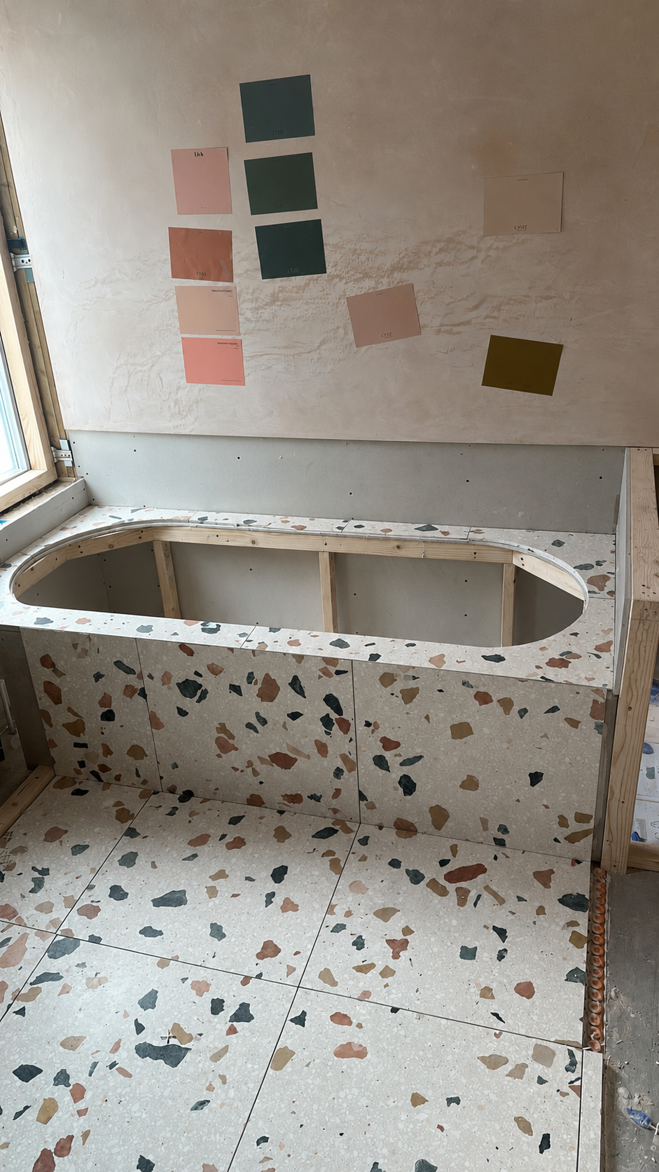 Under construction bathroom with a terrazzo tub surround and tiled floor, with color sample swatches on the wall.