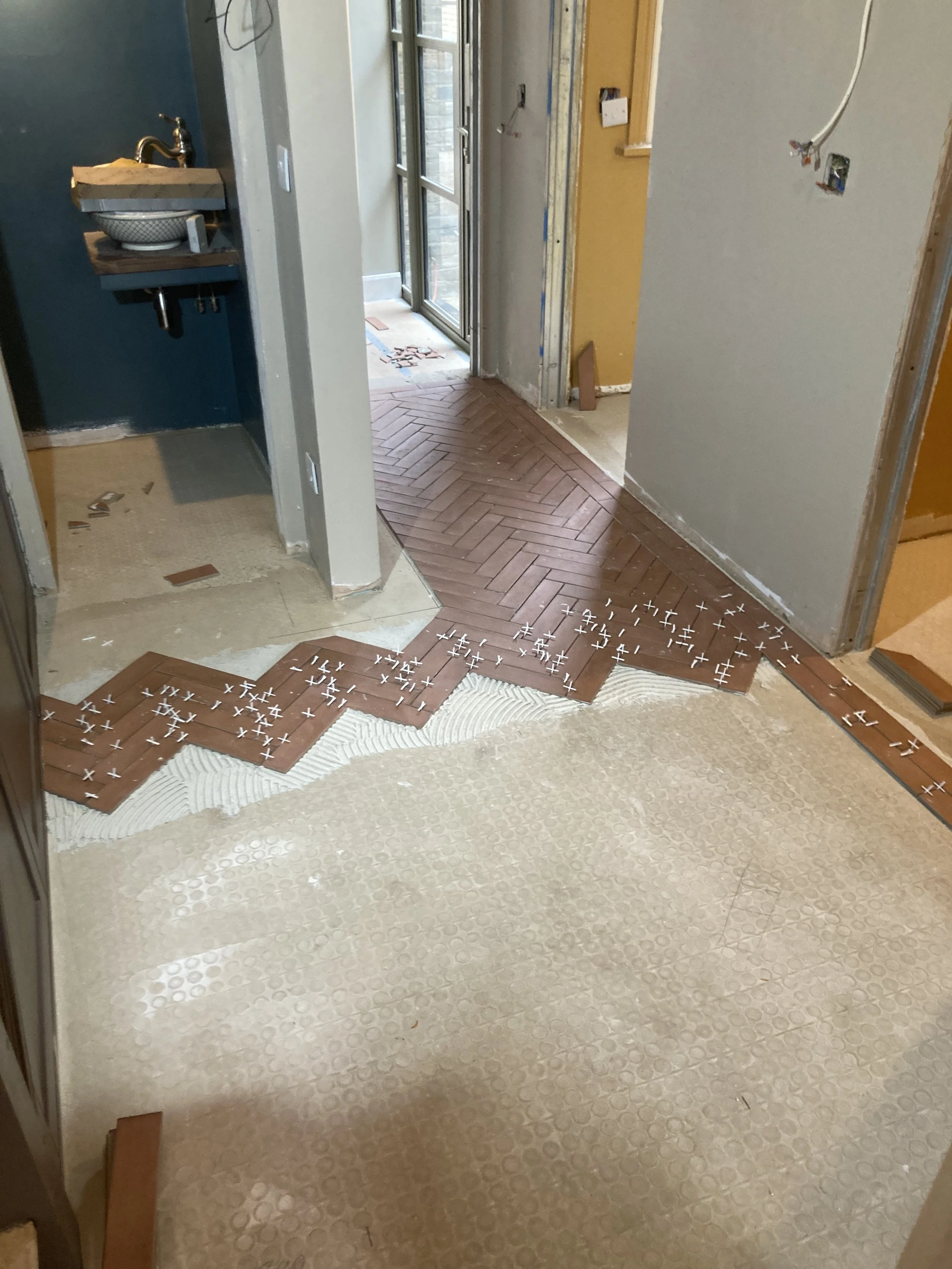 The image shows a room under renovation with partially installed herringbone wooden flooring. There is a step or threshold with underlayment materials visible at the bottom of the image. To the left, a small sink is being installed, attached to a sma