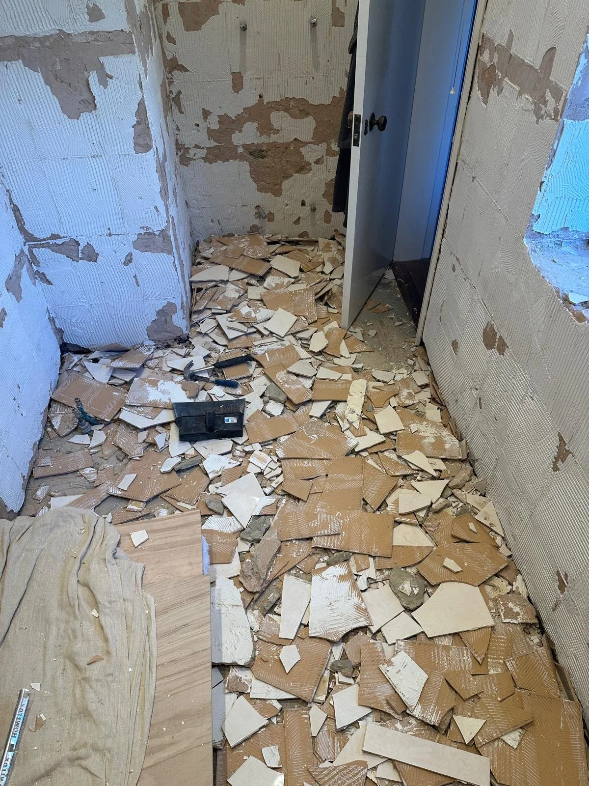 A room under renovation with broken tiles scattered across the floor and exposed drywall on the walls. An open door reveals a closet or small space with hanging clothes, and construction tools are on the ground.