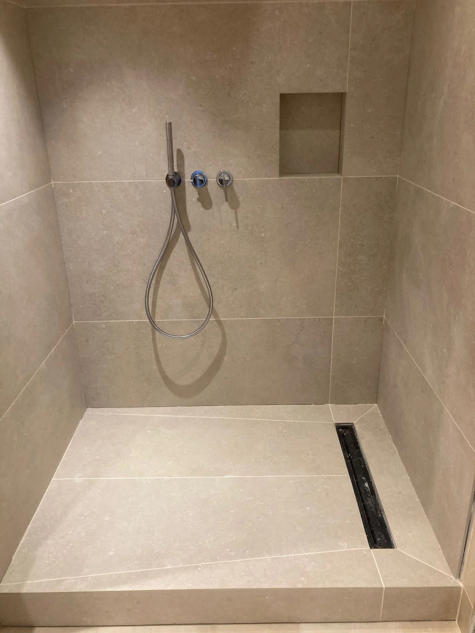 A minimalist shower with beige tiled walls and floor, a stainless steel handheld showerhead, a wall-mounted faucet, a built-in wall niche, and a linear floor drain.