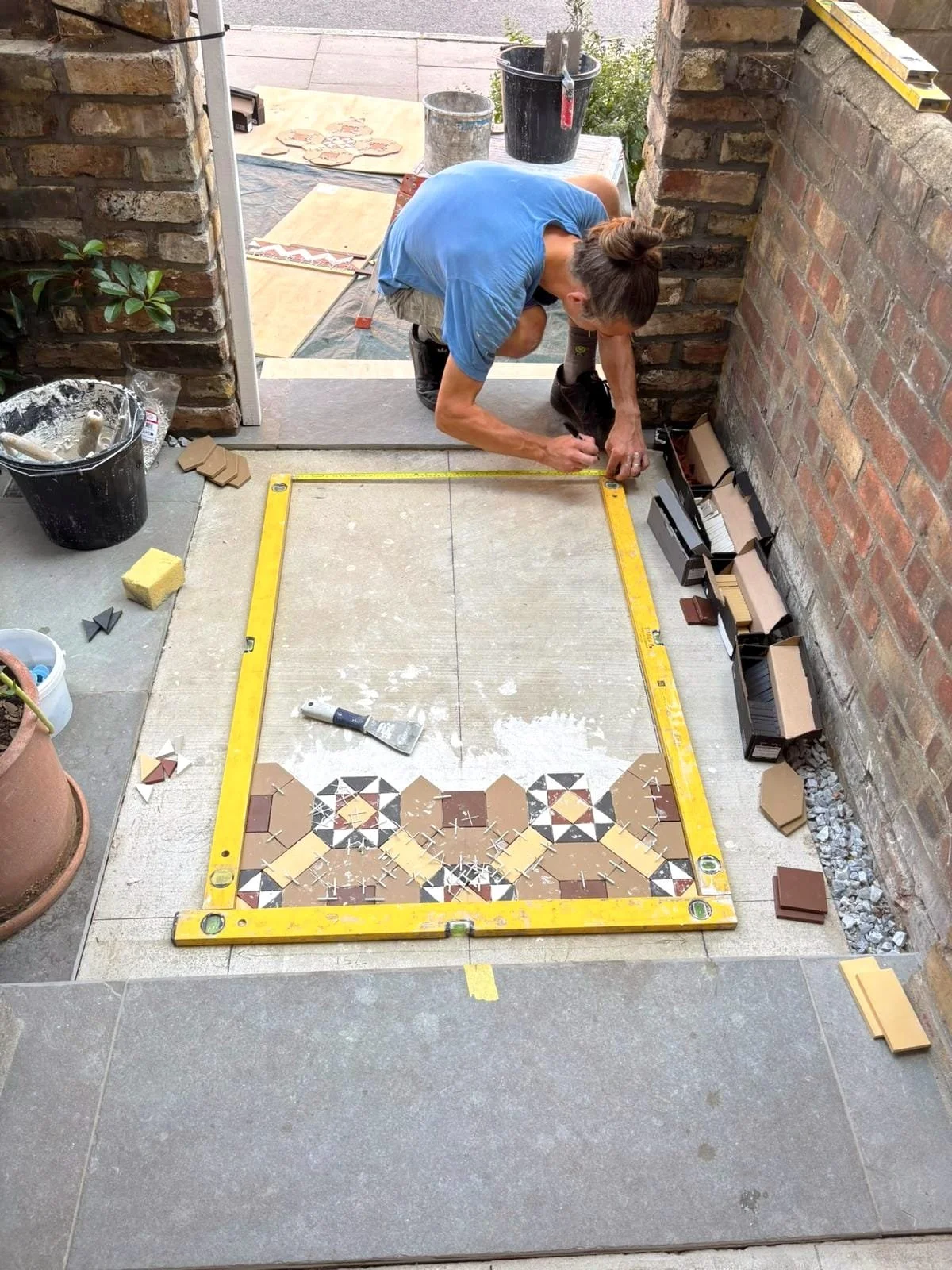 Person installing decorative ceramic tiles on a floor using a measuring tool, with tile supplies and tools around.
