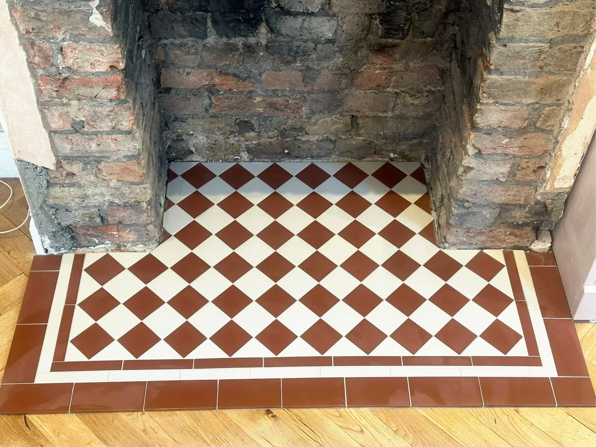 Part of an interior wall made of exposed bricks and a firebox with a tiled hearth. The hearth has a checkered pattern of white and brown tiles, with a border of larger brown tiles and an inner border of white tiles.