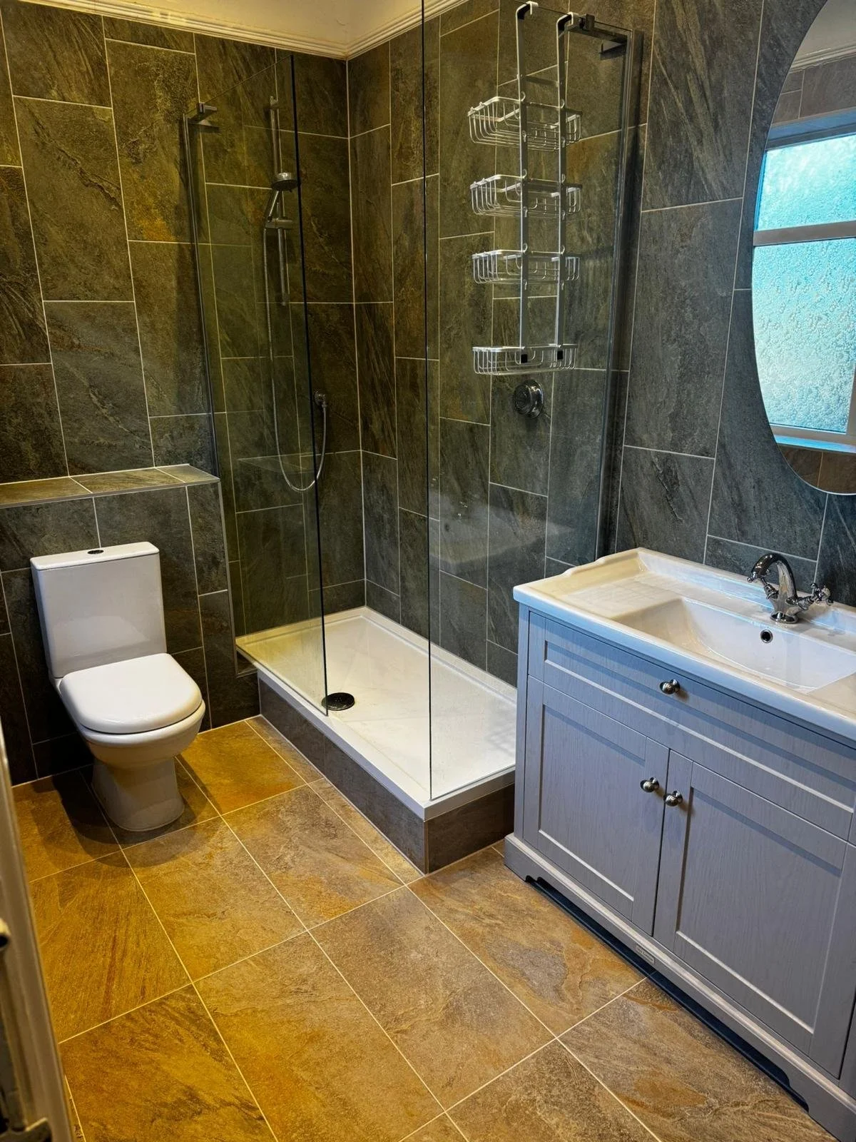 Bathroom with dark brown tiled walls and a matching tiled floor. Features a white toilet on the left, a walk-in shower with glass doors and a white shower tray in the center, a metal shower caddy hanging inside the shower, and a light gray vanity with a sink and chrome faucet on the right. A window with frosted glass is behind the vanity.