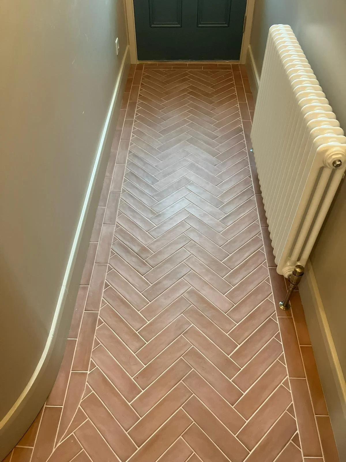 Indoor hallway with herringbone patterned ceramic tile floor, beige walls, black door at the end, white radiator on the right, and baseboard trim along the walls.