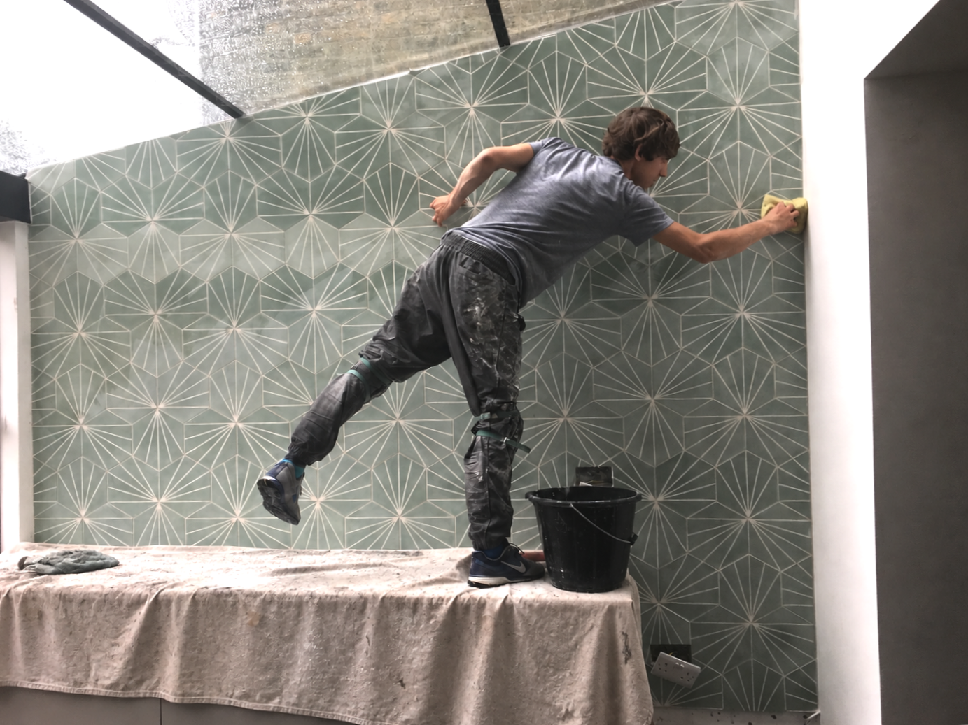 A man painting a geometric patterned wall while standing on a table covered with a beige cloth. He is using a sponge or brush to apply paint to the wall near the ceiling, with a bucket of black paint on the table. The room has a modern, minimalistic design.