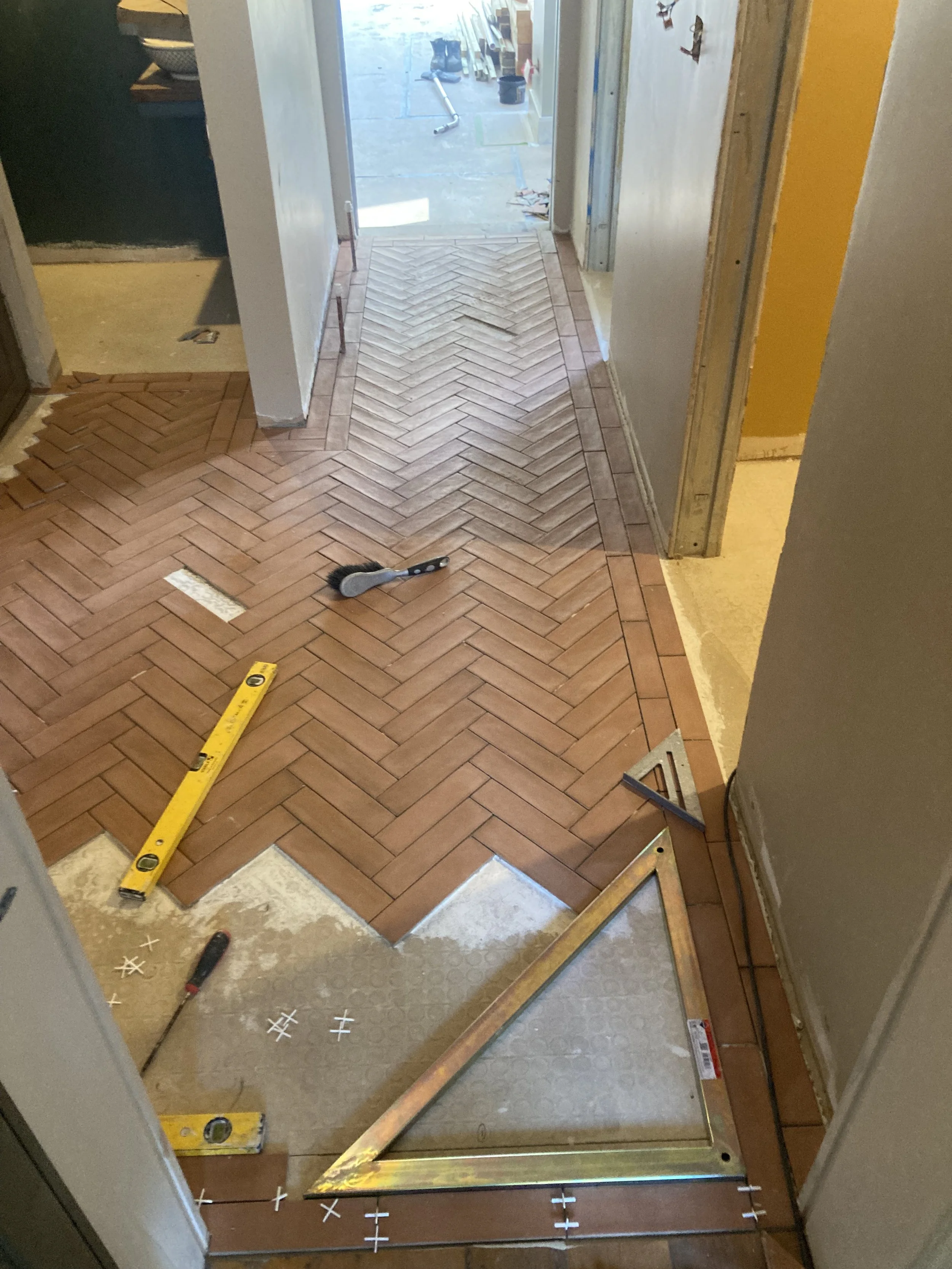 A hallway under renovation with herringbone-patterned wooden flooring being installed. Tools such as a level, a brush, and other construction equipment are visible. The hallway leads to an outdoor area with construction materials and a pipe on the gr