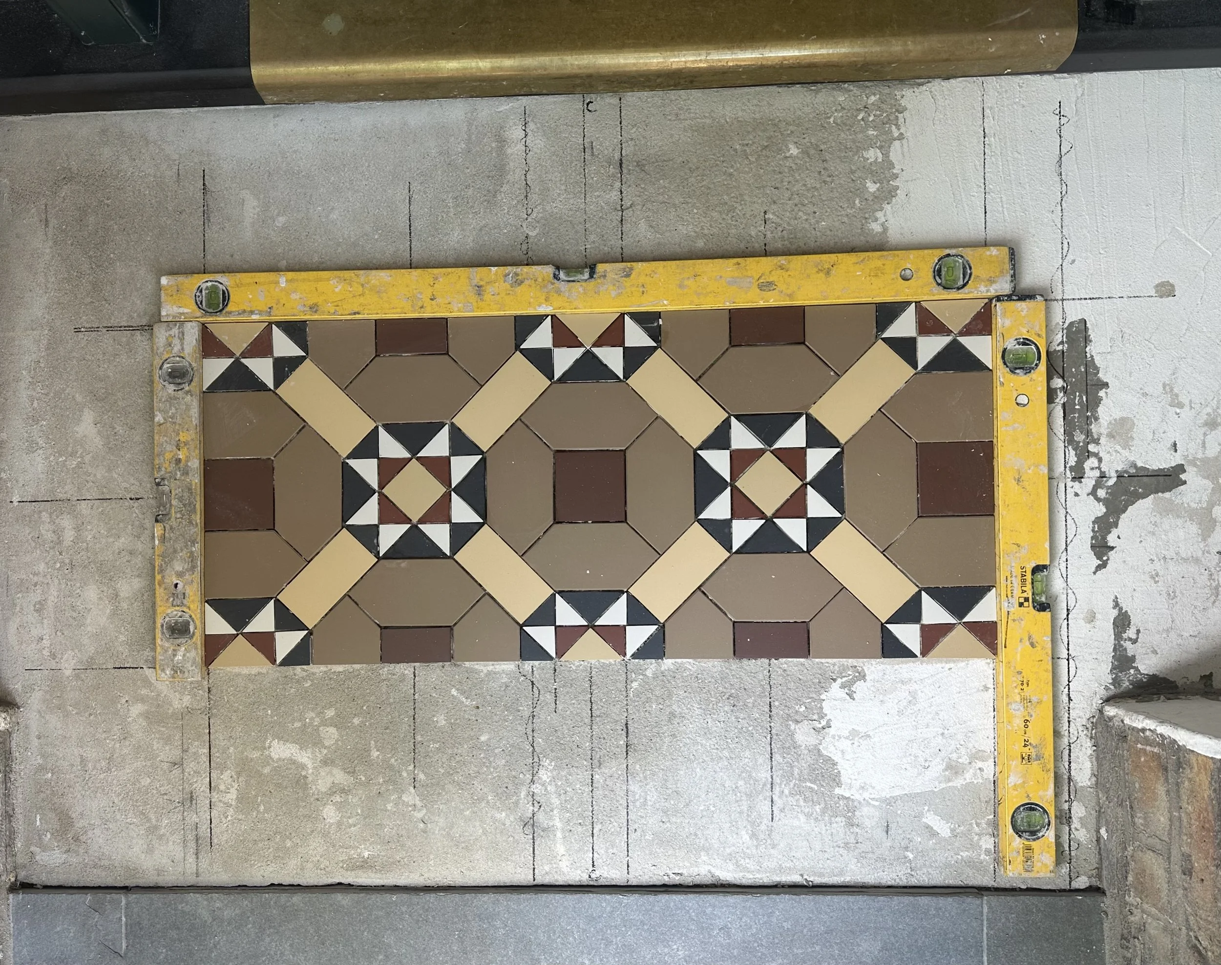 A patterned tile section on a construction floor, bordered by a yellow level tool and a cement surface.
