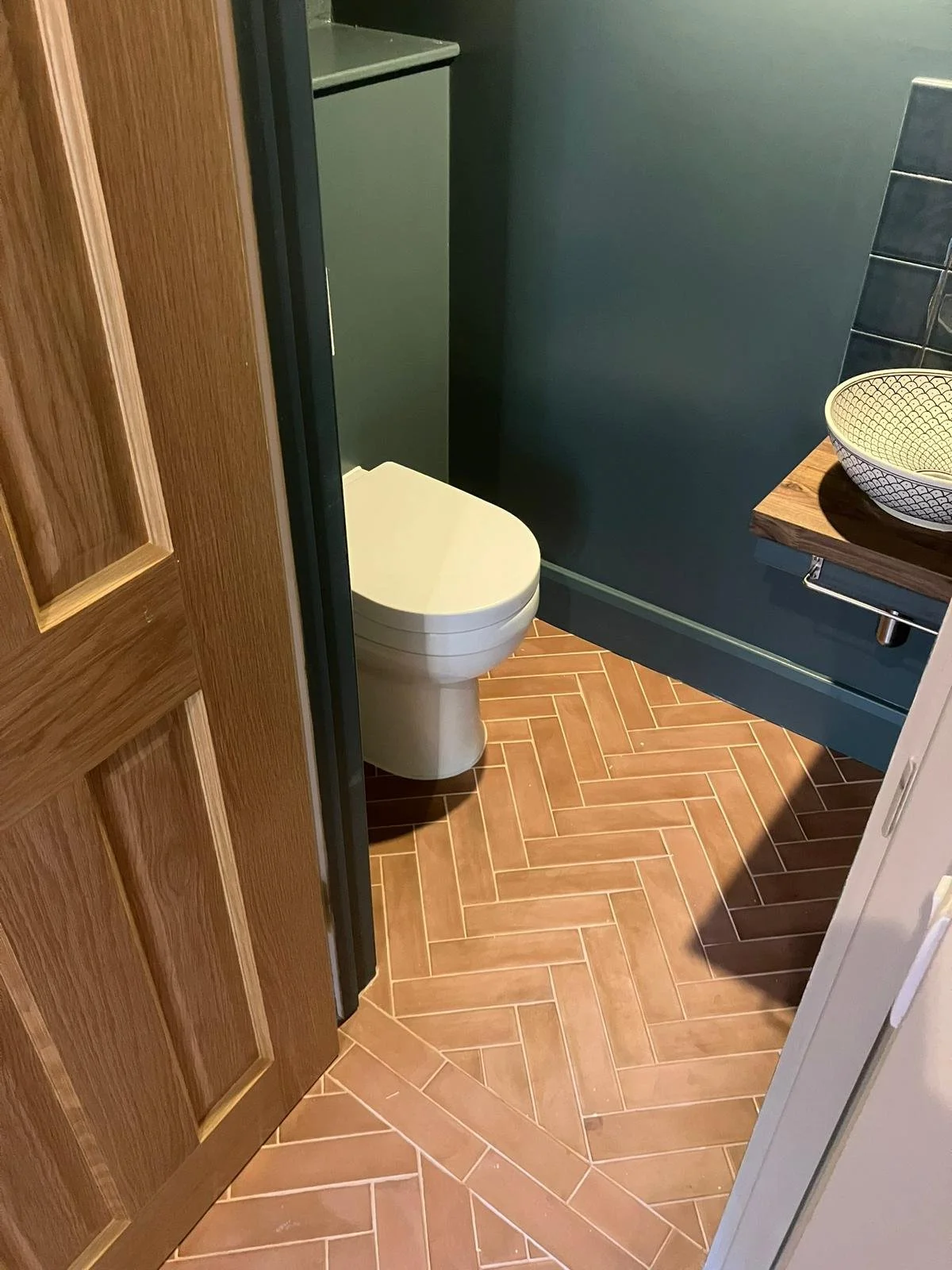 Small bathroom with a closed toilet, a wooden door, a dark teal wall, a wooden edge shelf with a patterned bowl, and herringbone-patterned terracotta floor tiles.