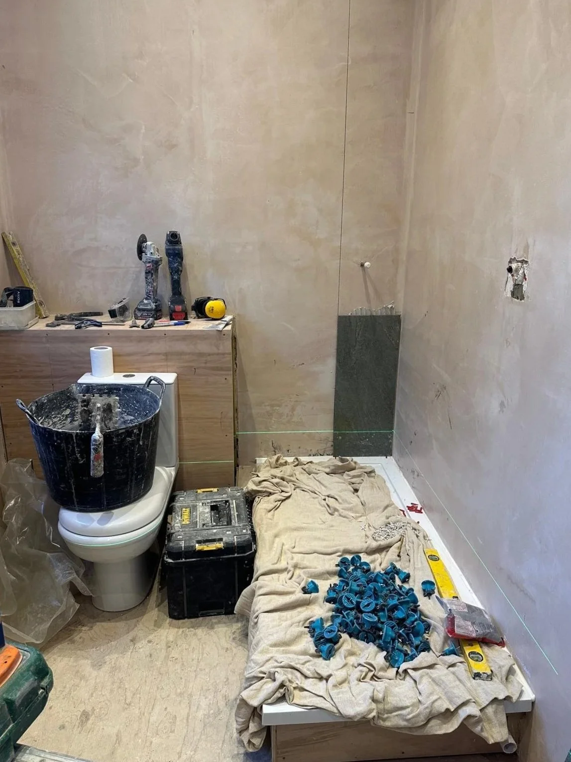 A bathroom under construction with tools, a toilet with a bucket on top, a fitted wooden cabinet, construction materials, and a cloth covering the floor and a bed of small blue plastic pipe fittings.