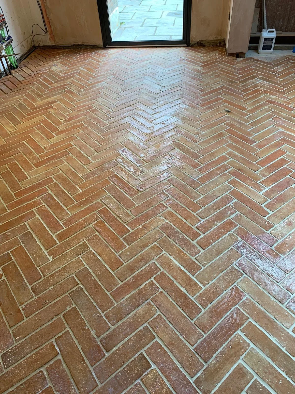 A newly installed herringbone brick floor in a room with a sliding glass door leading outside. Construction materials and tools are partially visible.