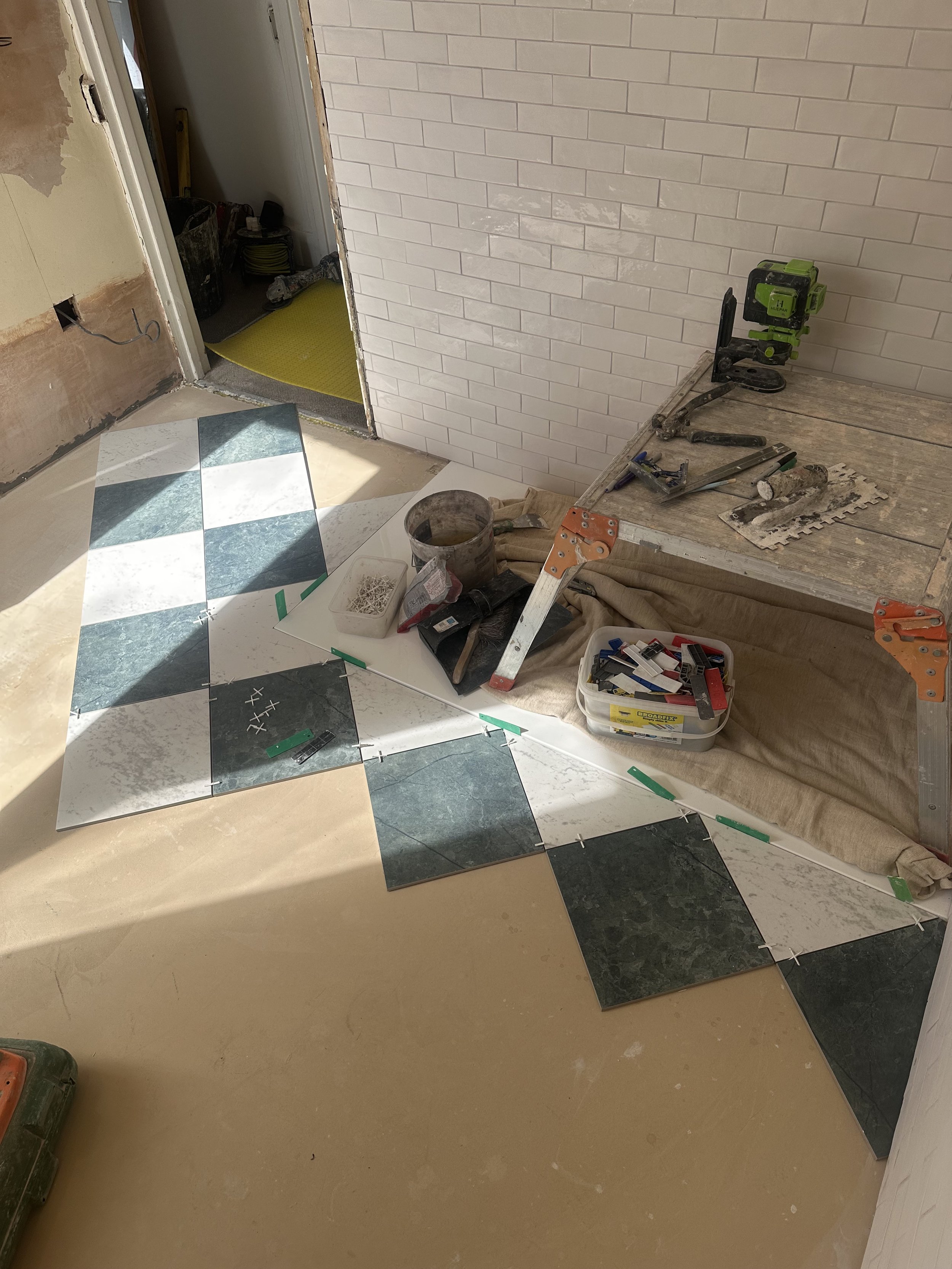 A room under renovation with a partially tiled wall, some tools and construction materials, a step ladder, and a section of floor with black, white, and beige tiles. The doorway has exposed edges and construction debris on the floor.