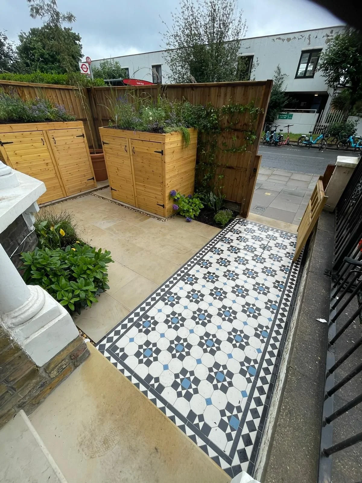 A front yard with patterned black and white tiles, beige stone tiles, a wooden fence, green plants, and a view of bicycles and a building across the street.