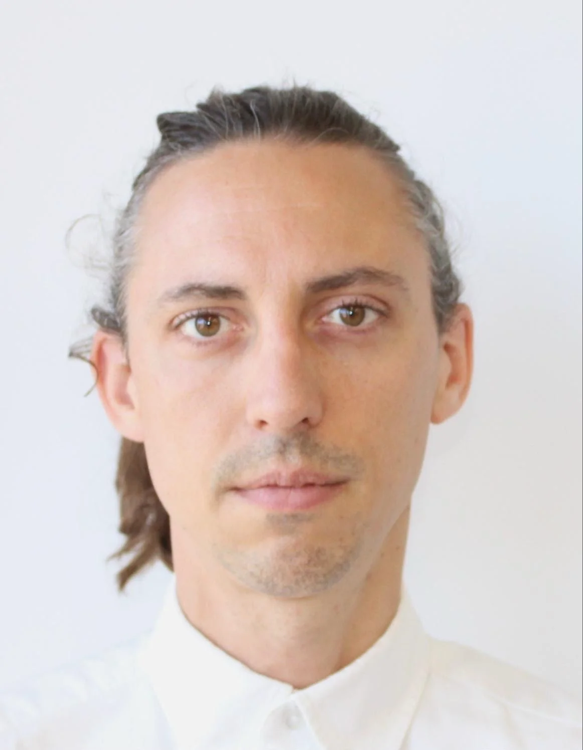 Portrait of a man with long brown hair, light skin, wearing a white collared shirt, looking directly at the camera with a neutral expression.
