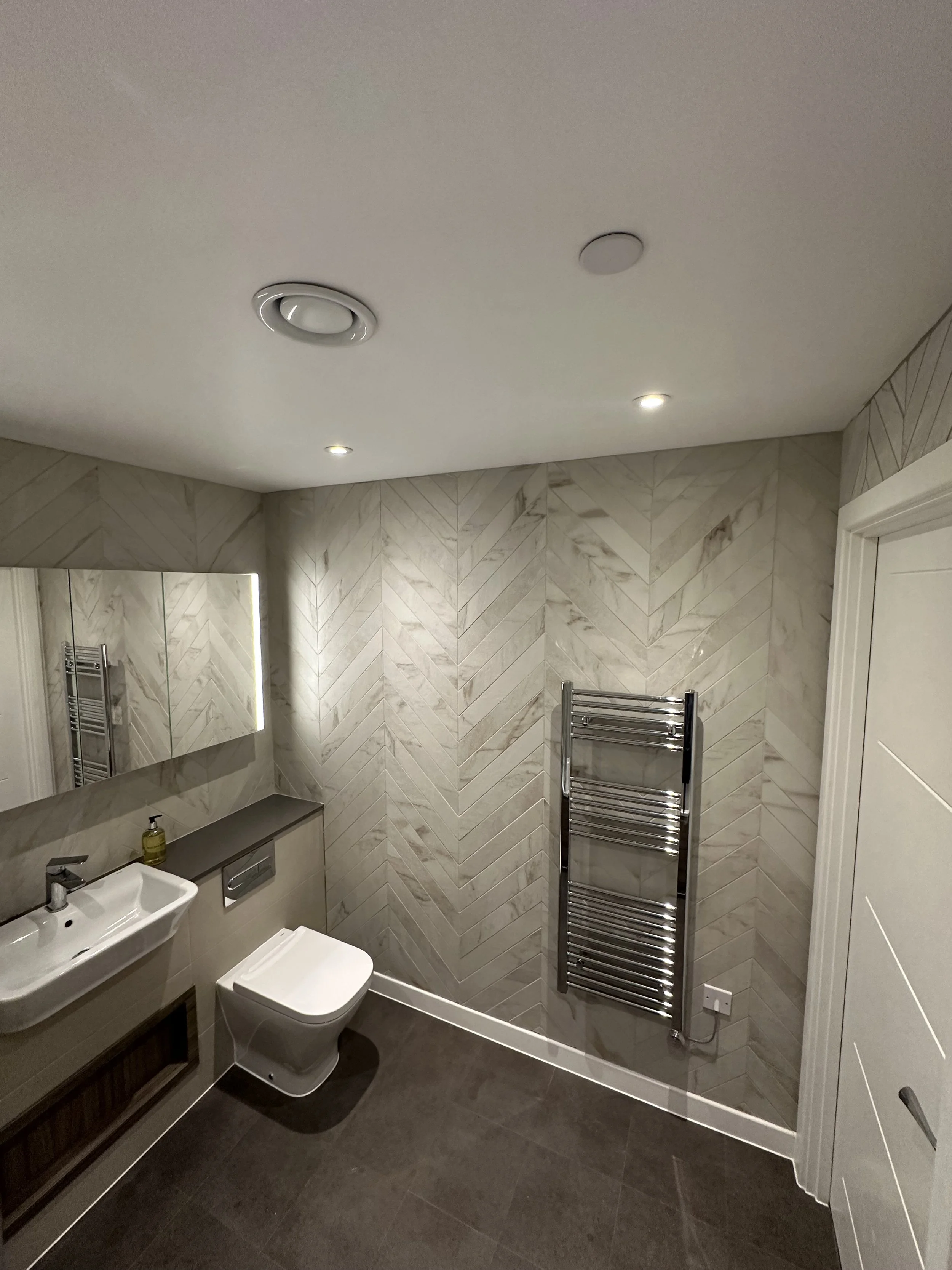A modern bathroom with a white toilet, a sink with a mirror, a towel radiator, and beige marble-patterned wall tiles.