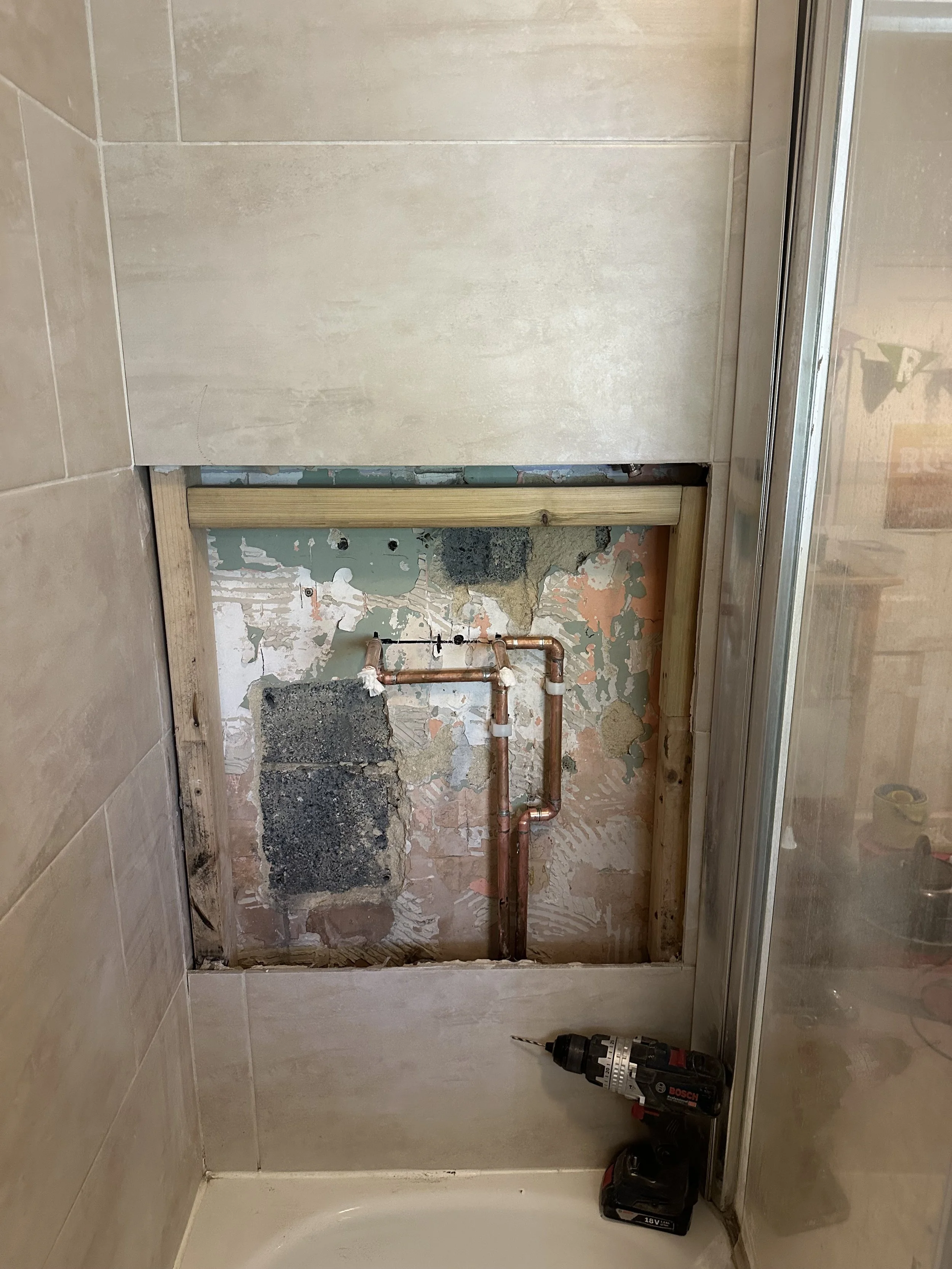 Bathroom renovation in progress with exposed plumbing and wall damage, a Bosch cordless drill on the floor, and a partially removed shower door.