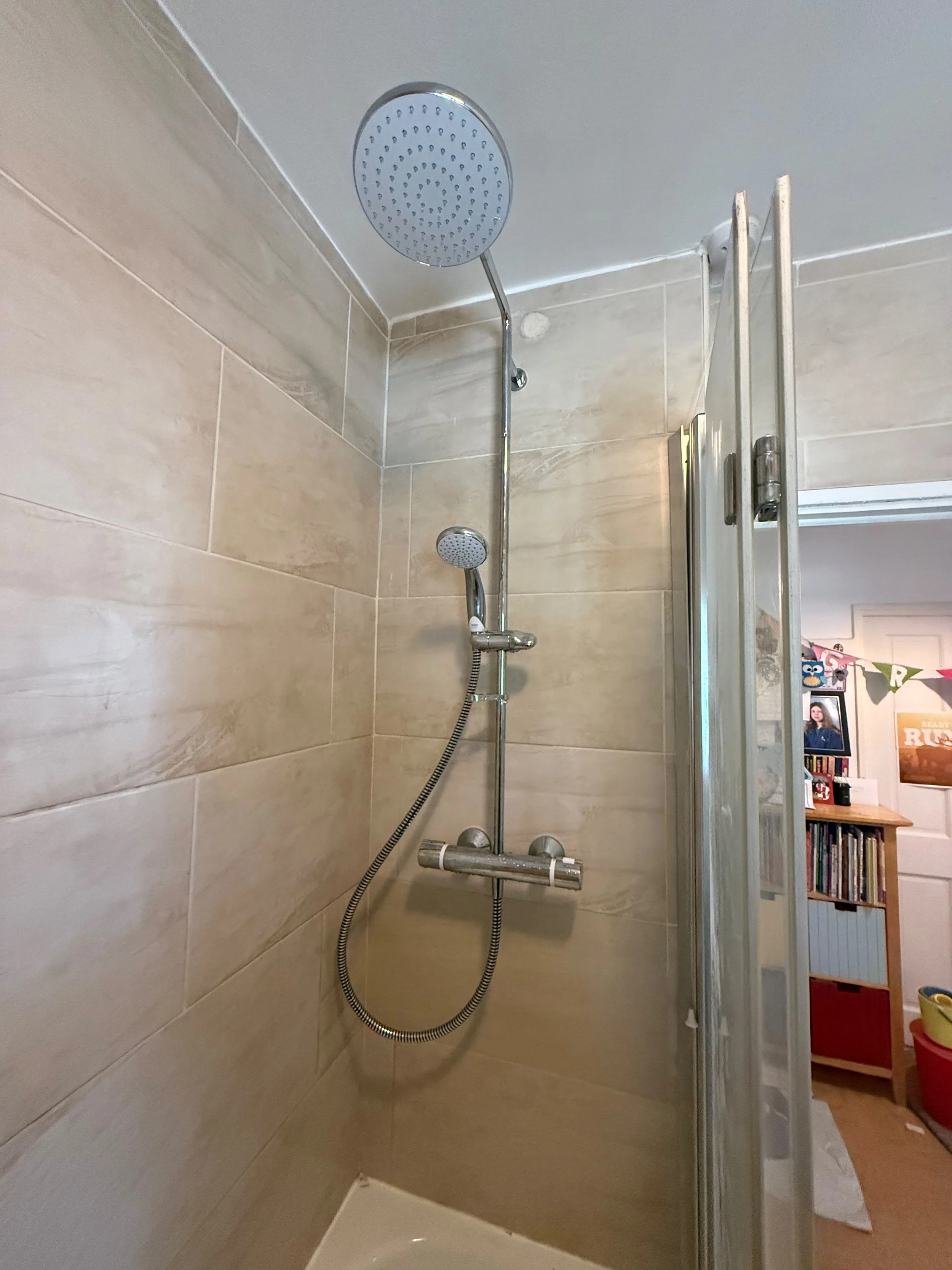 Shower with beige tiled walls, a round overhead shower head, a handheld shower head, and a sliding glass door, with part of a room visible in the background.