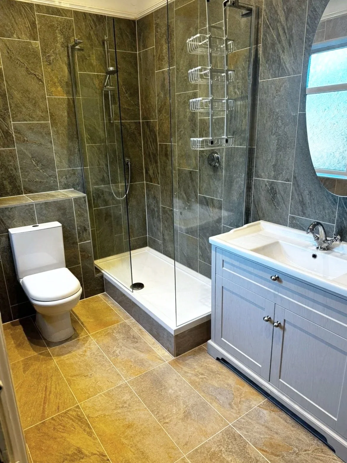Bathroom with a white toilet, a glass shower enclosure with a showerhead and shelf, a sink with a light gray vanity, a mirror, a small window, and beige tiled flooring and walls.