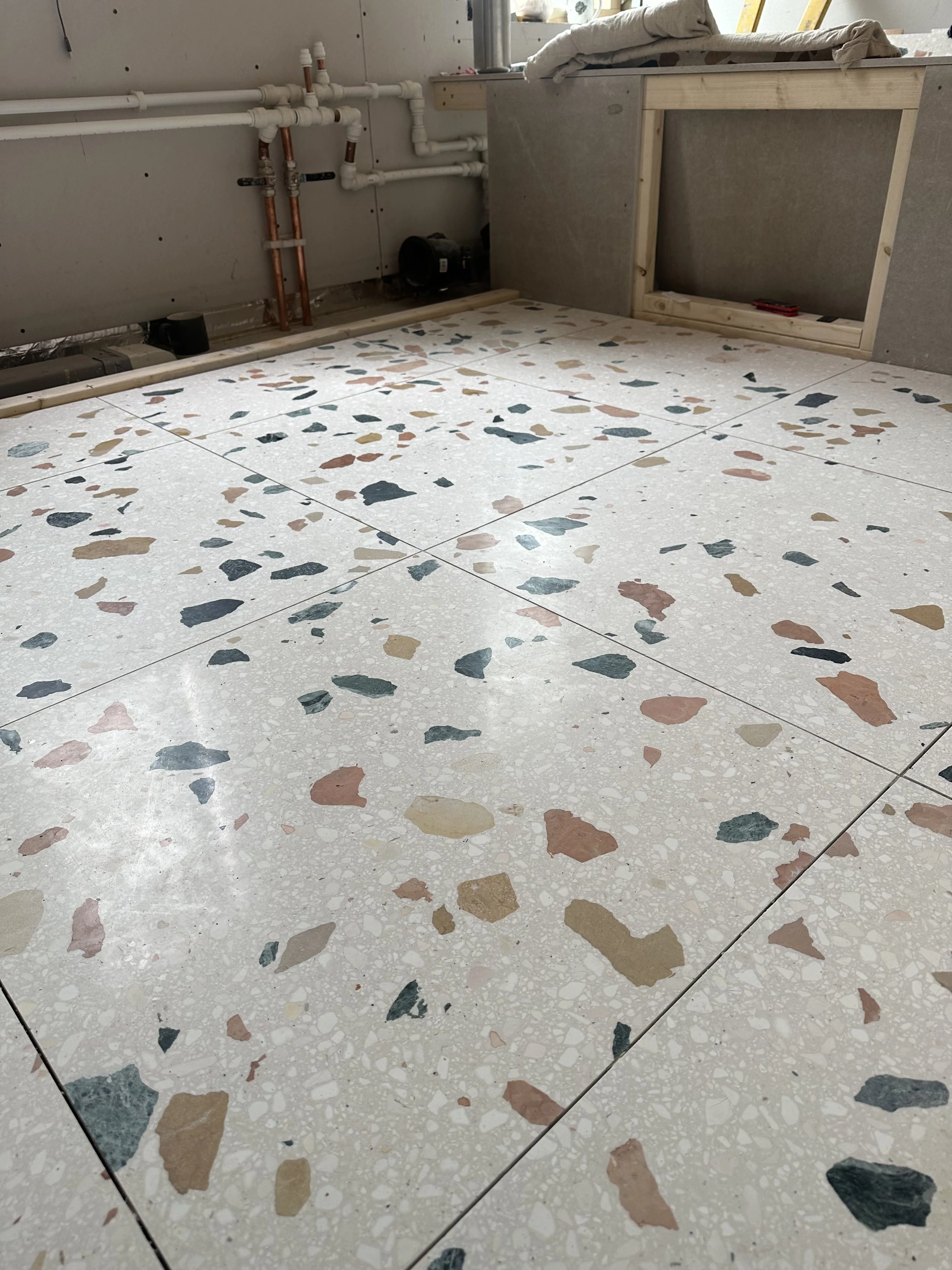 A floor with terrazzo tiles featuring multicolored stone fragments in shades of pink, black, beige, and blue, surrounded by unfinished construction with plumbing pipes and a partially built wall.