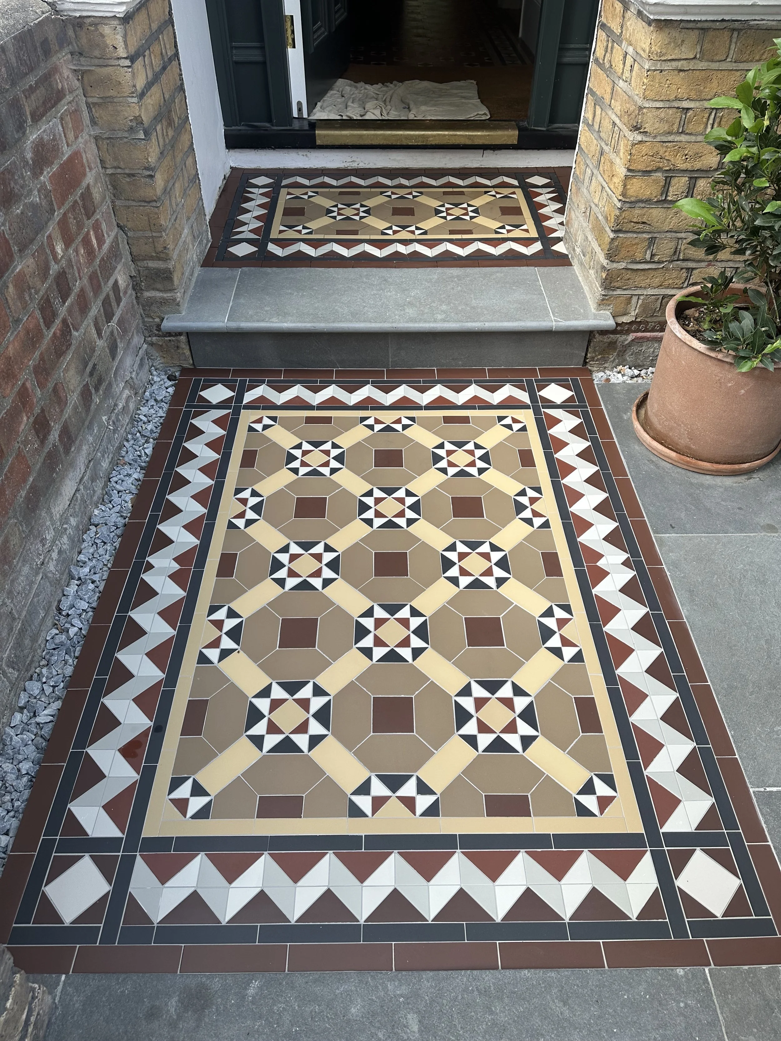 View of an ornate tiled entrance mat and doorstep with brick walls and potted plant to the sides, leading into a dark interior.