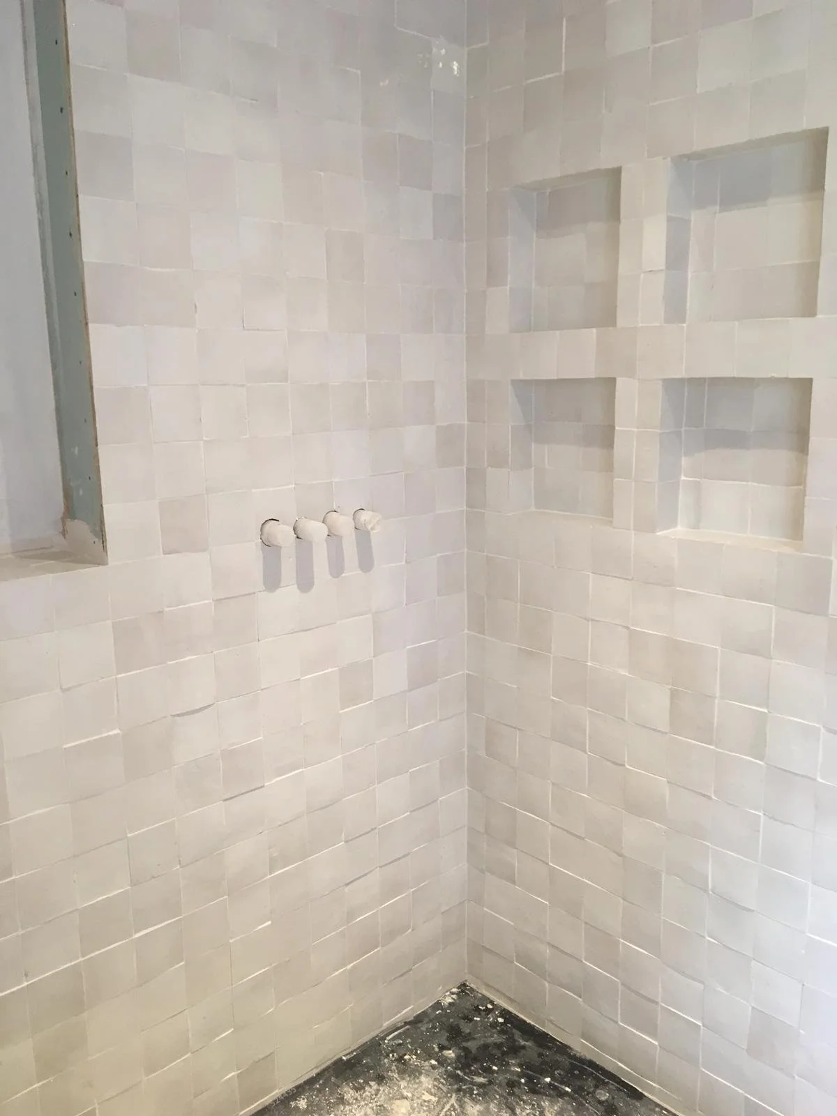 A corner of a bathroom shower area with newly installed beige tiles, built-in wall shelves, and pipes for a shower fixture. The floor appears to be unfinished.