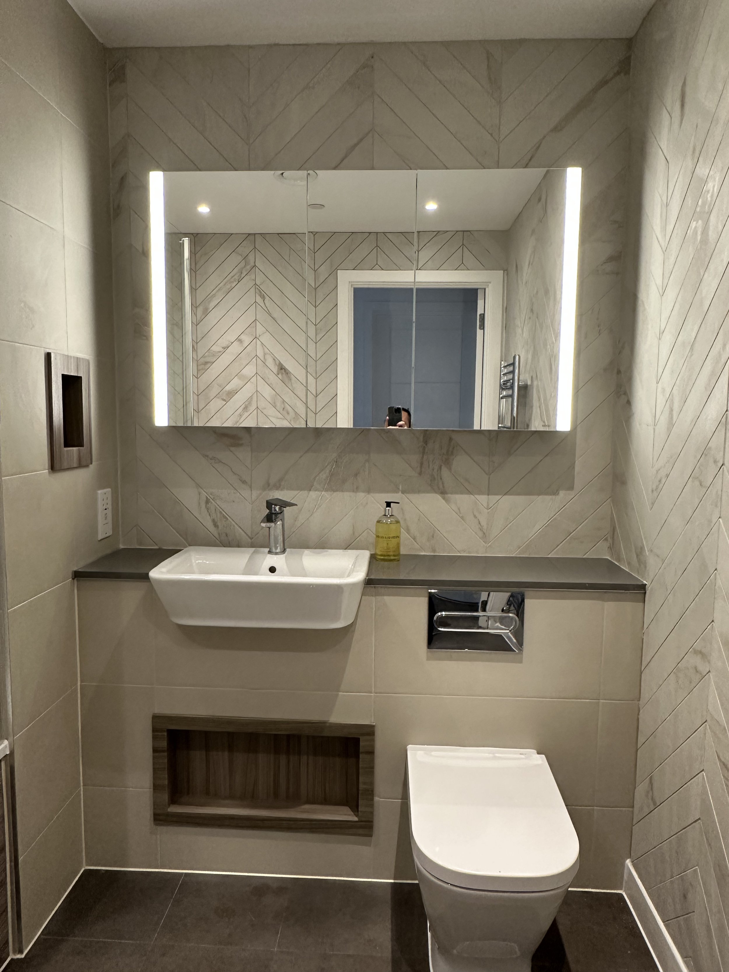 Modern bathroom with beige tiled walls, a white wall-mounted sink, a mirror with integrated lighting, a wooden shelf beneath the mirror, and a toilet with a concealed cistern.