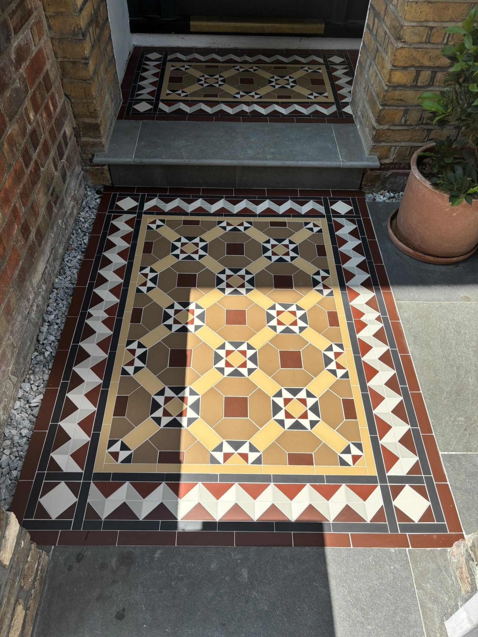 Decorative tiled porch entrance with patterned tiles on the floor, brick walls on either side, a potted plant to the right, and a step leading to a door.