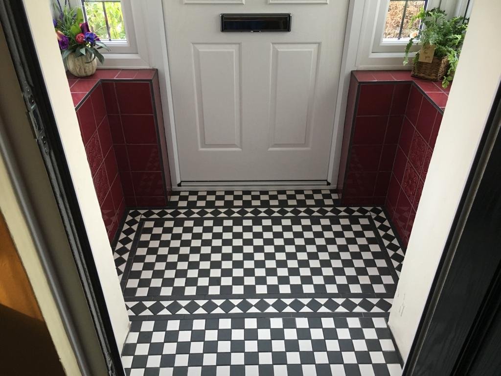 Entryway with black and white checkered tile floor, red tiled walls, a white door at the back, and windows on both sides with plants and flowers on the window sills.