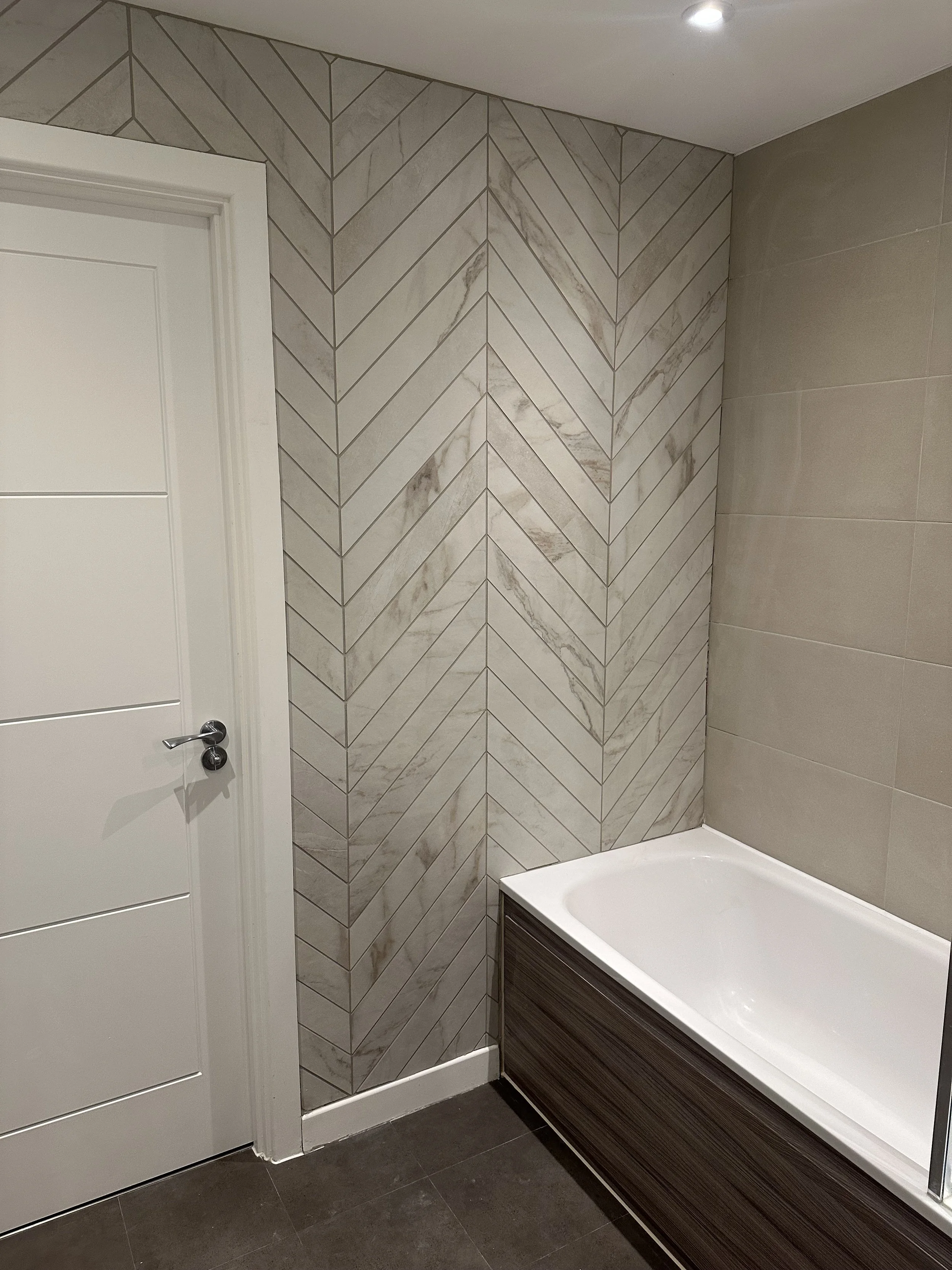 A bathroom corner showing a white door, marble patterned tile wall, beige tile wall, and a bathtub with dark wood paneling.